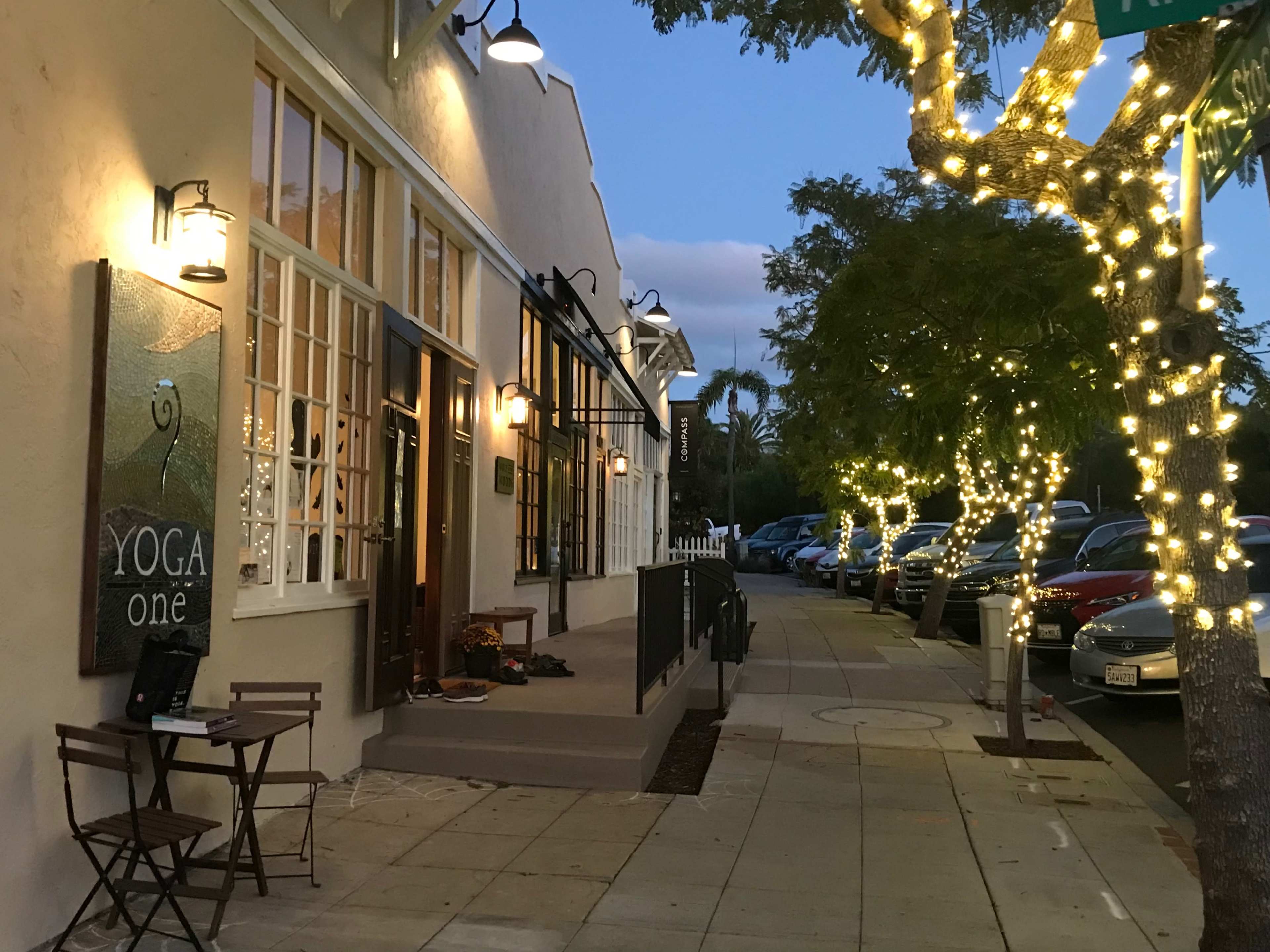 A yoga studio with string lights on trees is located along a sidewalk lined with parked cars in the evening.