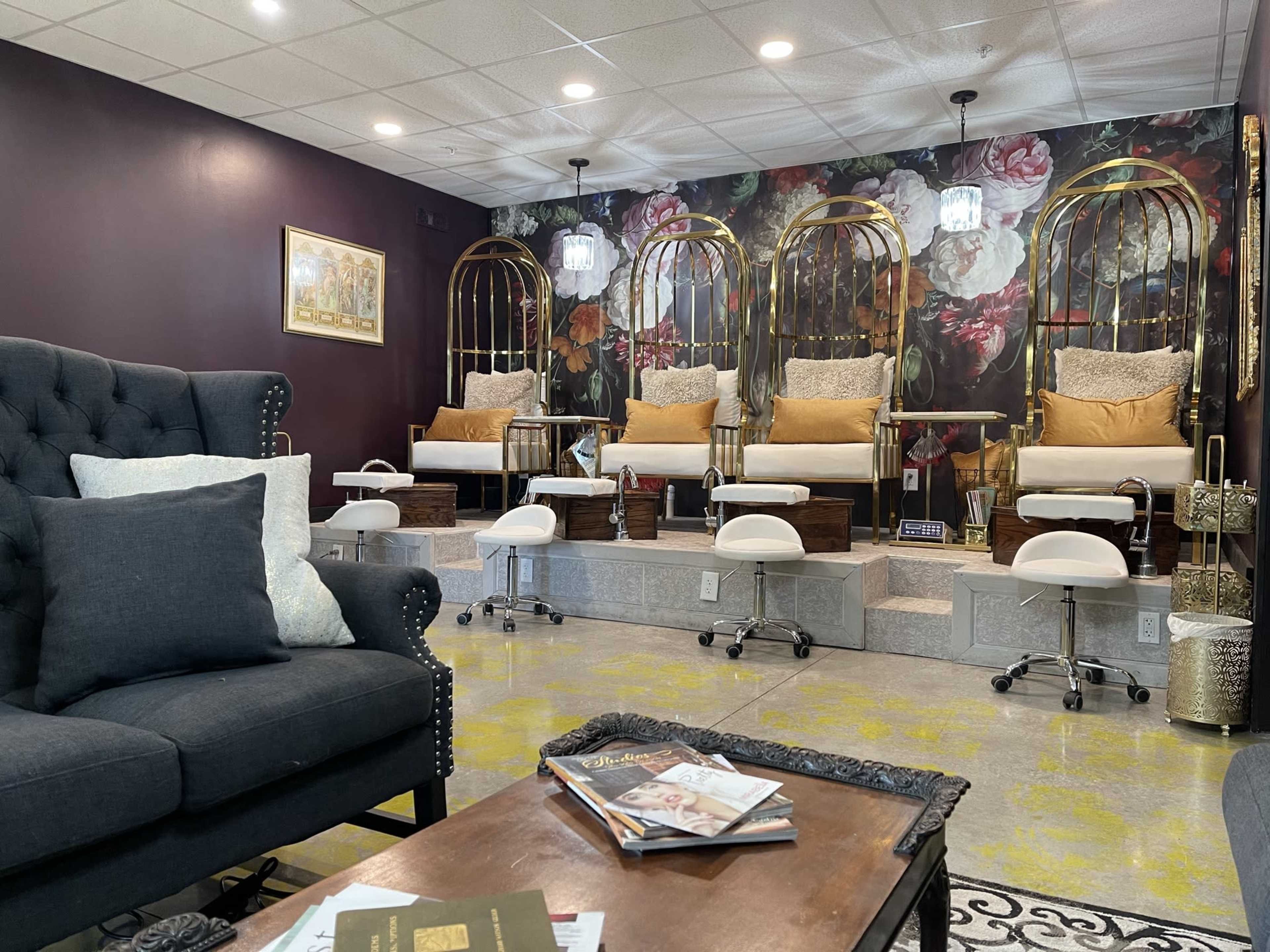A nail salon interior featuring four manicure stations with cushioned seating and decorative floral wallpaper in a dimly lit setting.