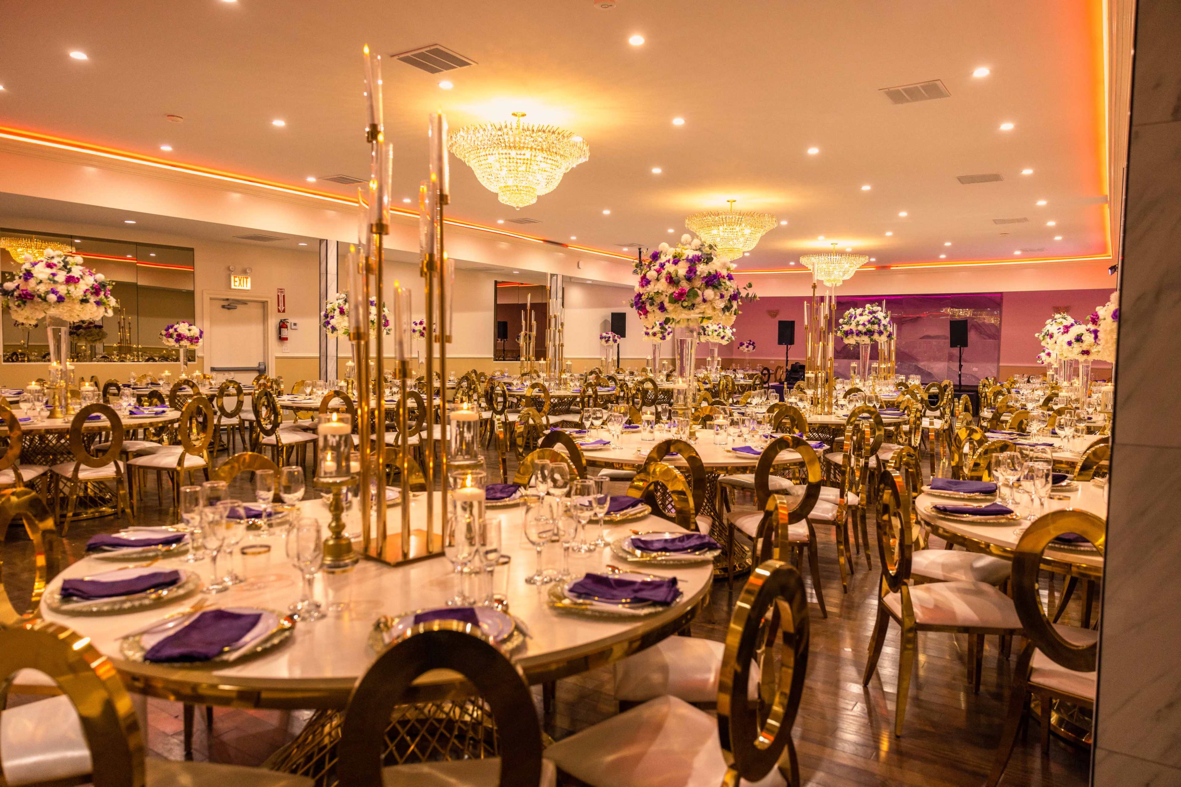A spacious banquet hall arranged for a formal event, featuring elegantly set tables with purple accents and floral centerpieces under bright chandeliers.