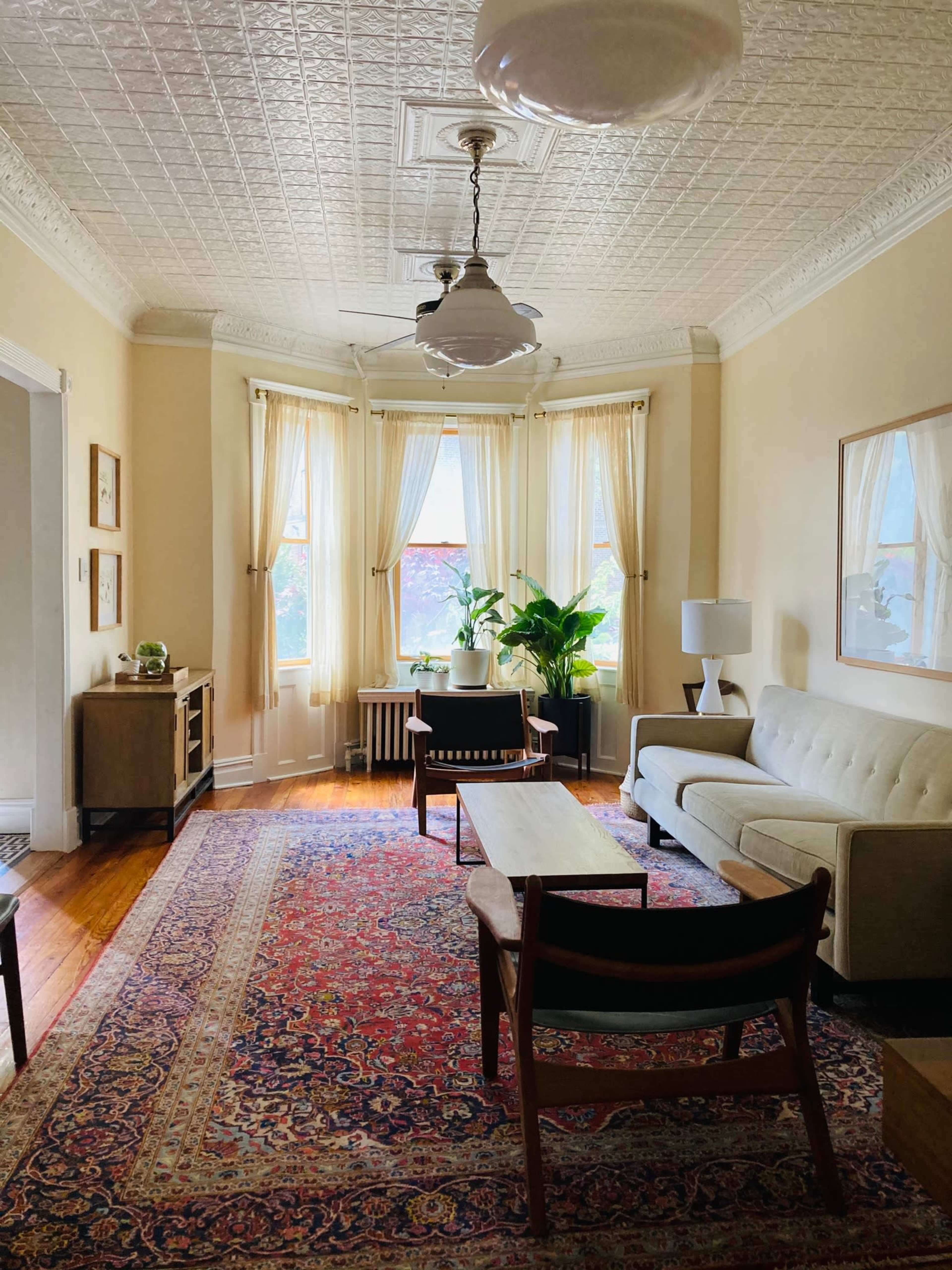 The image shows a living room with a sofa, coffee table, and a large window, adorned with curtains, and featuring plants and a decorative rug on the hardwood floor.