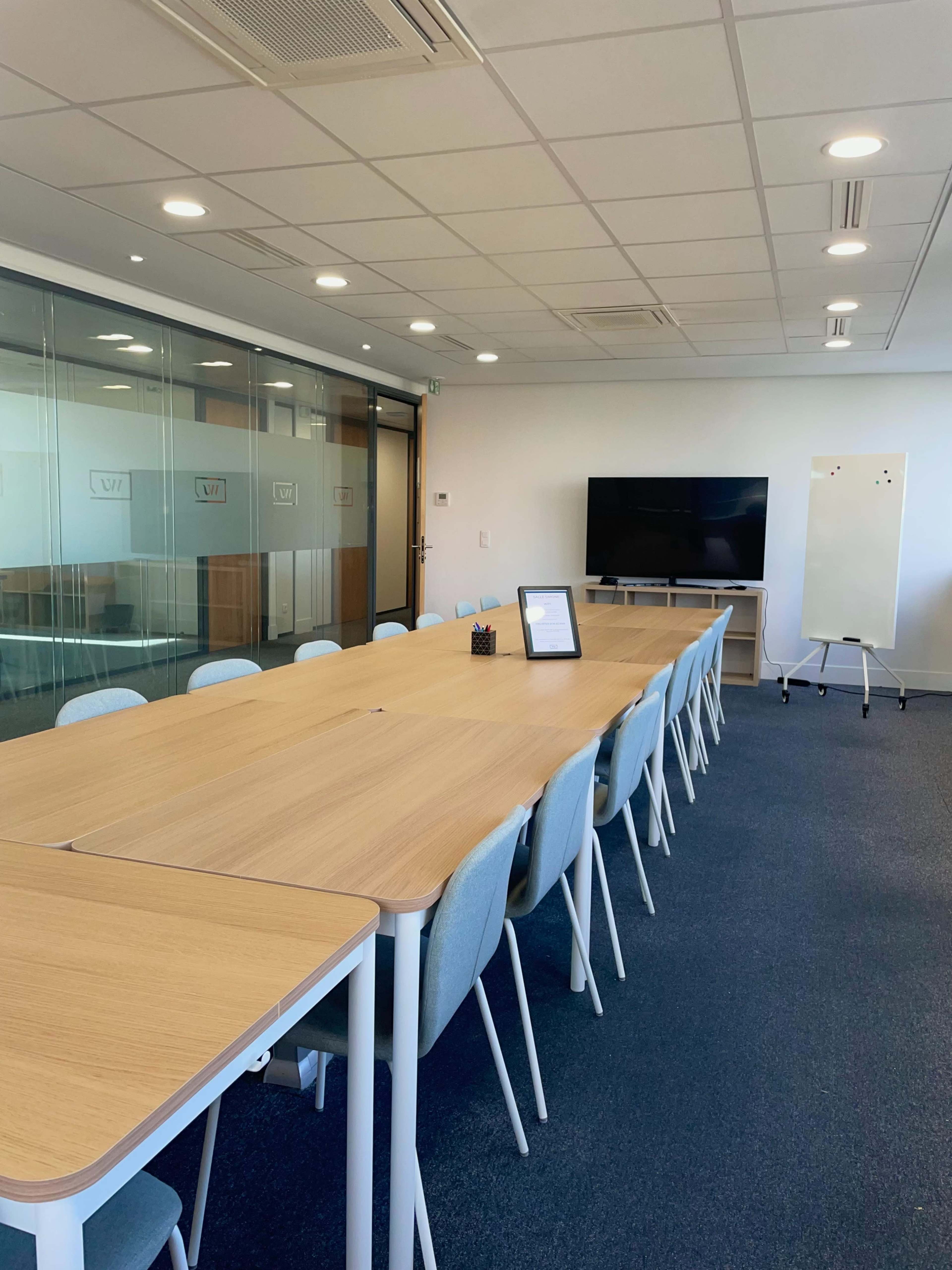 A modern conference room features a long wooden table with light blue chairs, a large screen on the wall, and a whiteboard nearby.