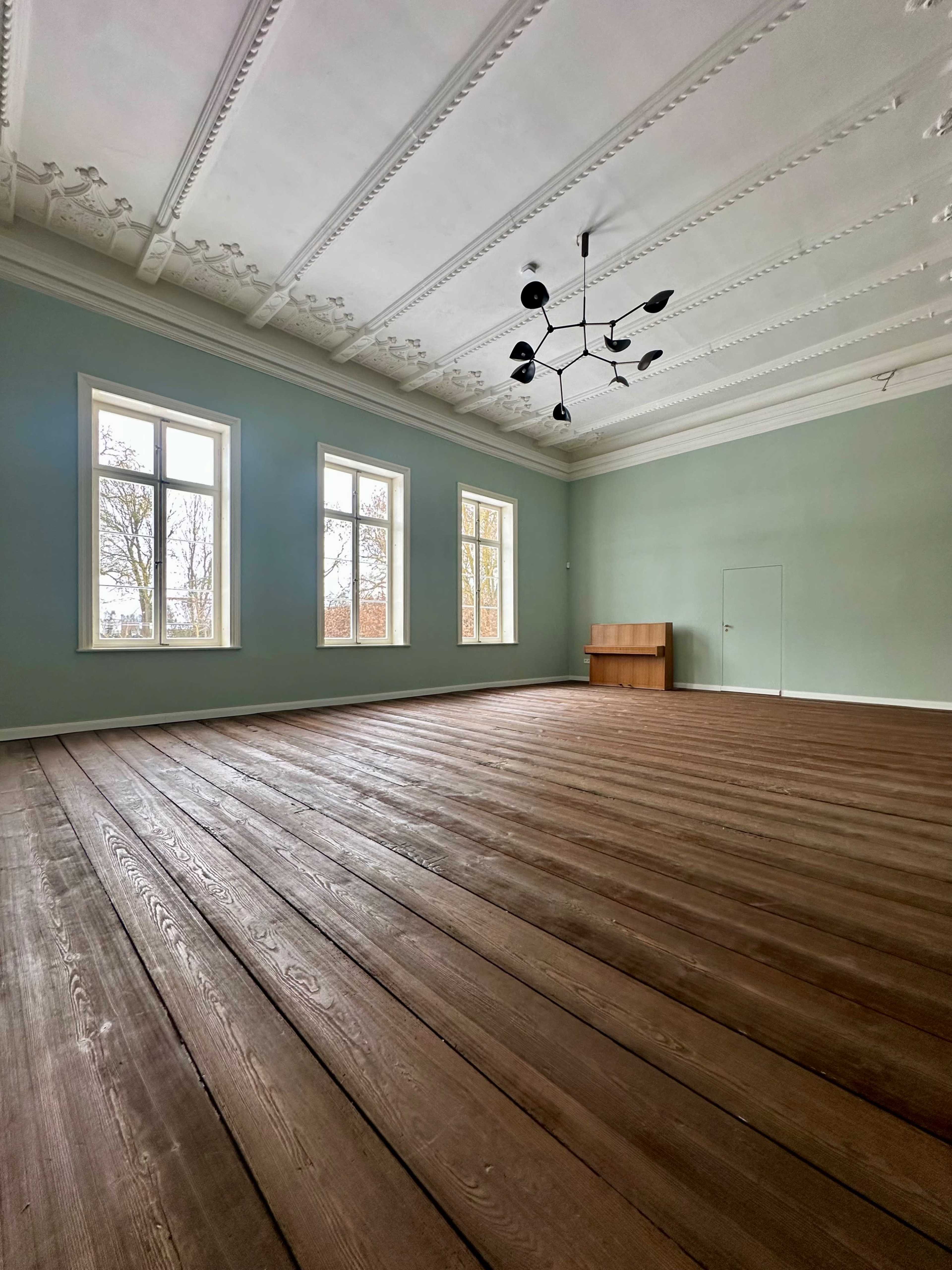 The image shows a spacious room with large windows, wooden flooring, and a minimalist light fixture.