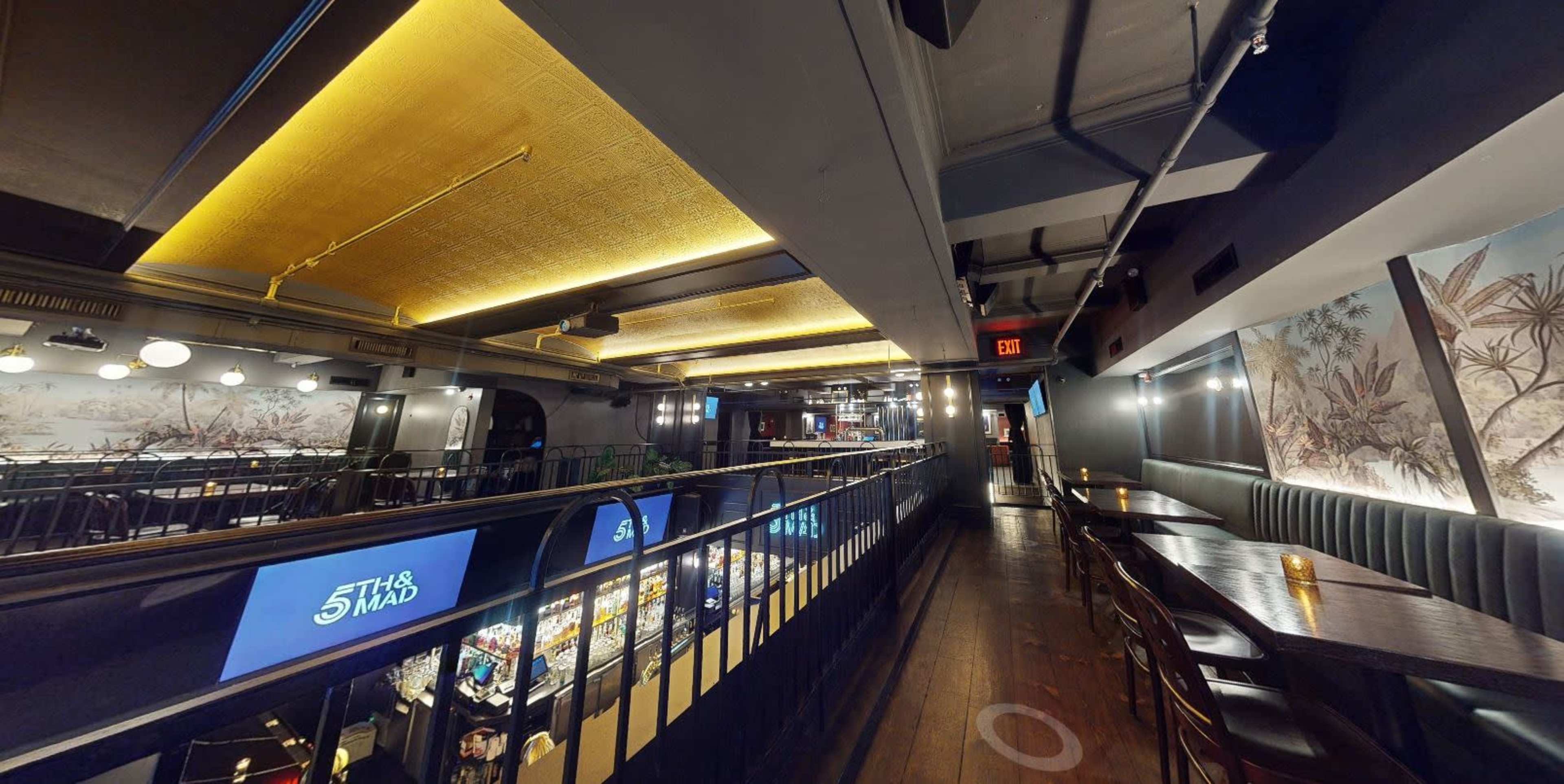The image shows the interior of a restaurant with a modern design, featuring wooden flooring, overhead lighting, and a balcony overlooking the dining area.