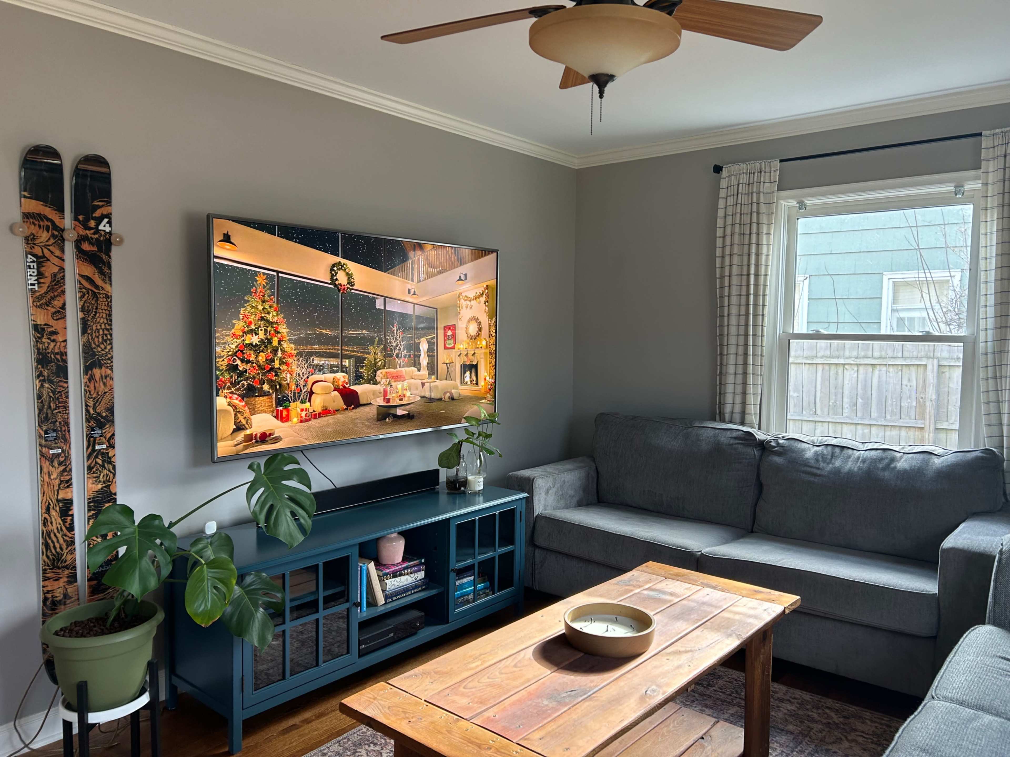 A living room features a gray couch, a wooden coffee table, a large TV displaying a festive scene, and ski equipment mounted on the wall.