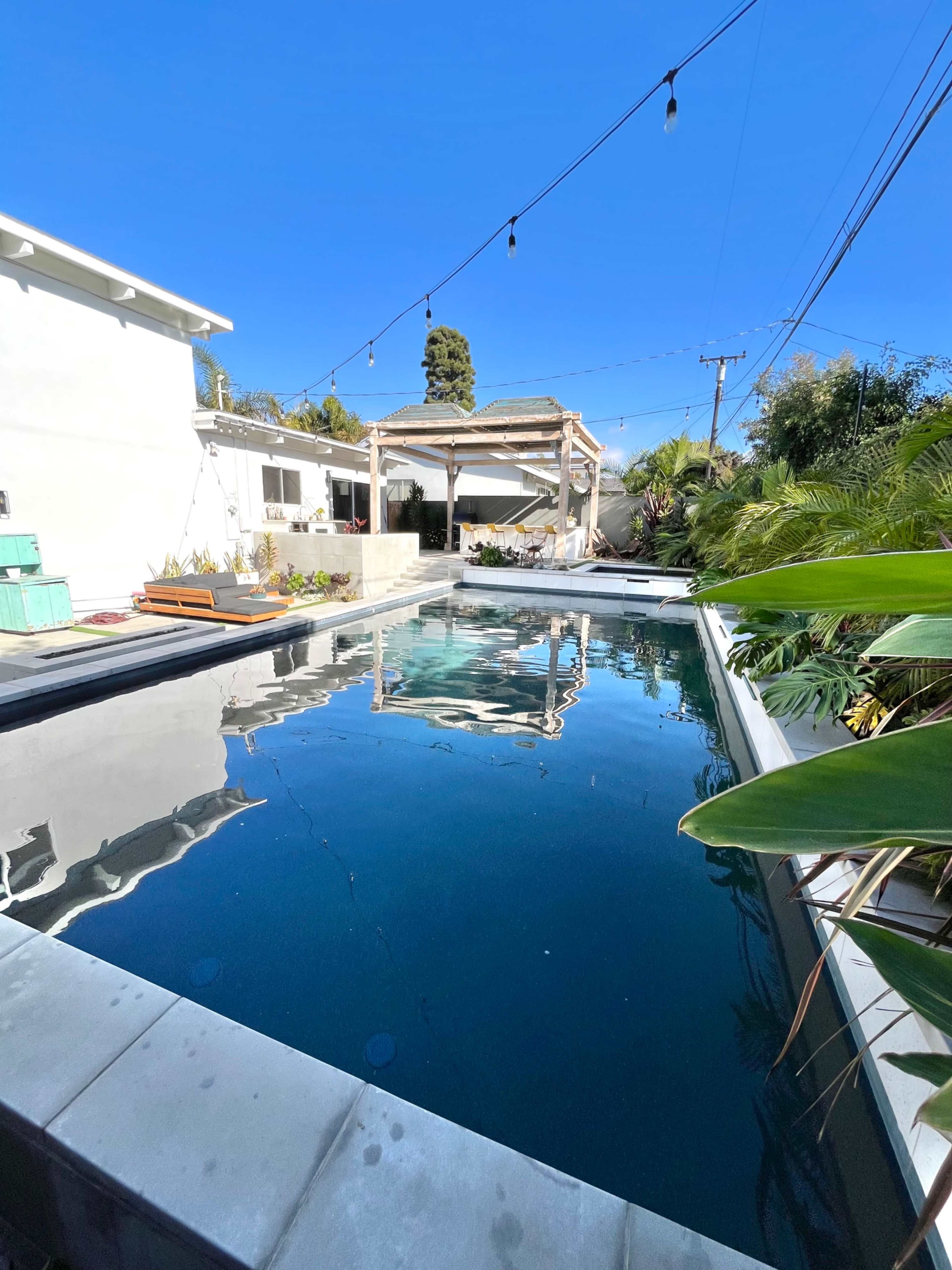The image shows a rectangular swimming pool surrounded by patio space and greenery under a clear blue sky.