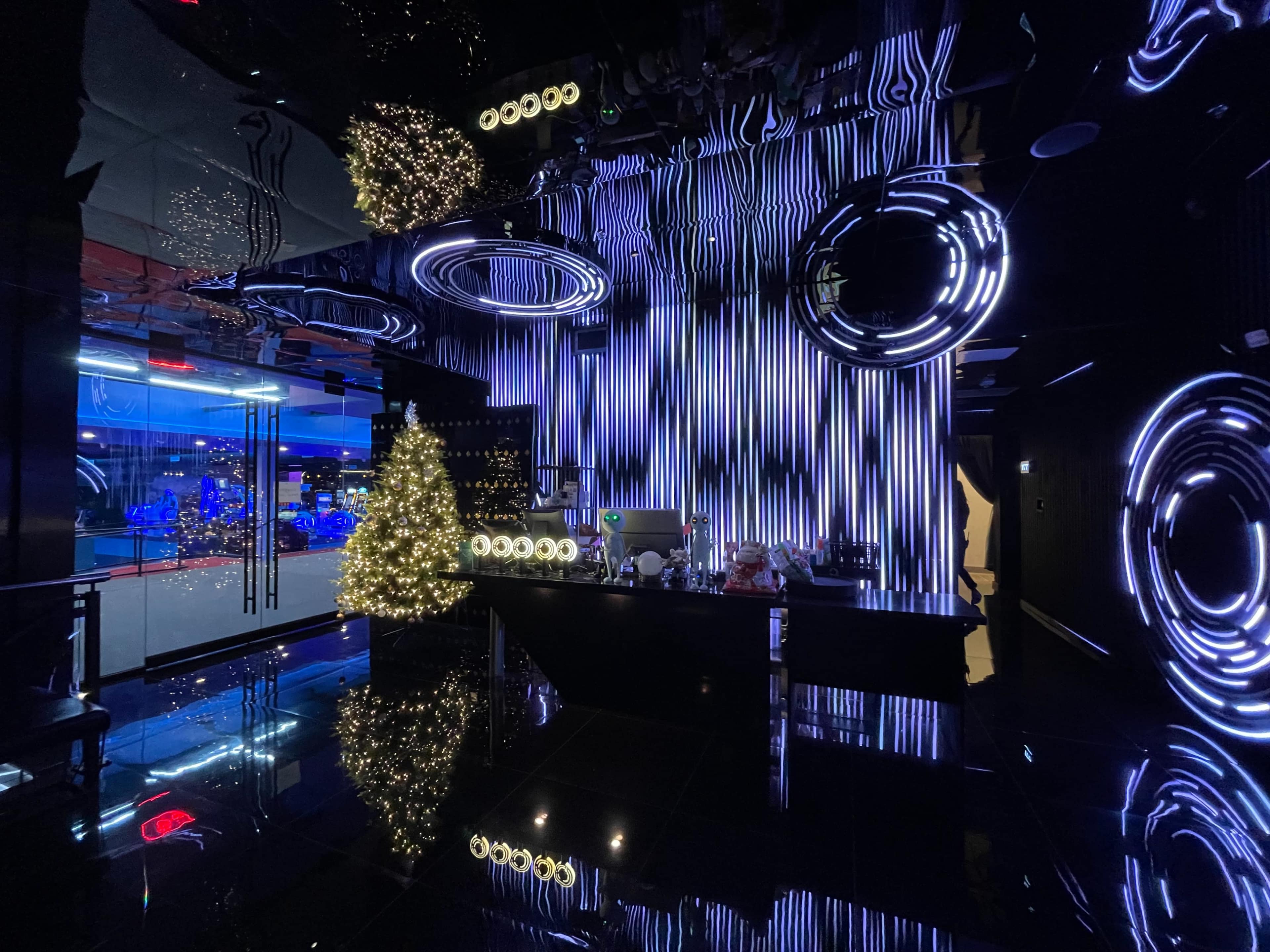A modern bar environment with a black reflective floor, striking neon lights, and a lit Christmas tree.