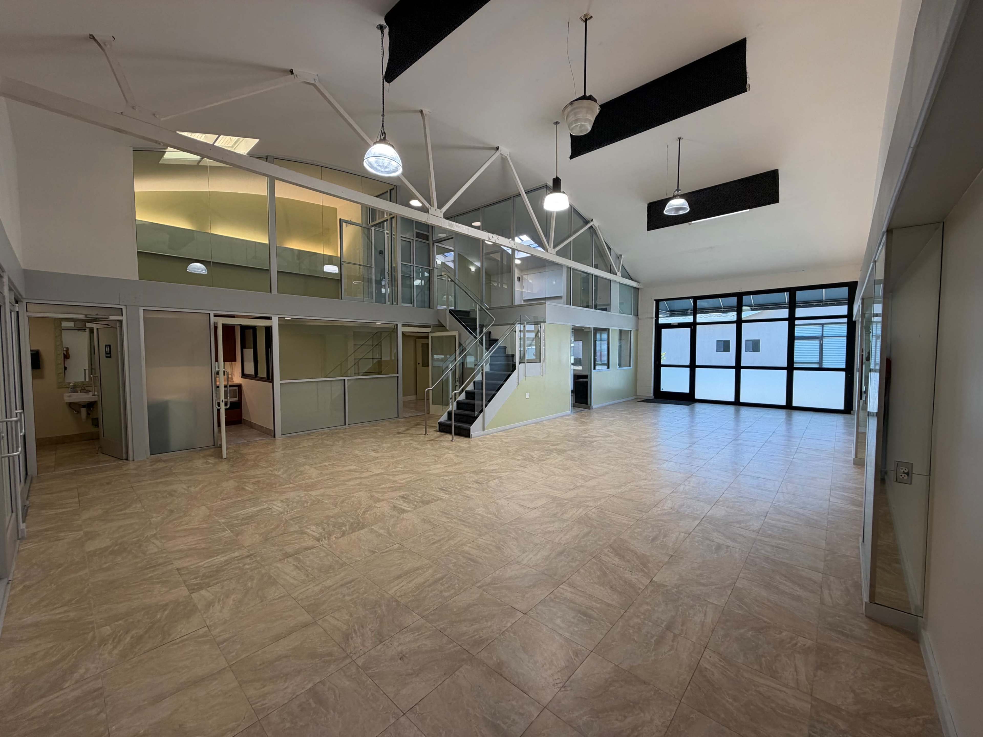 The image shows a spacious, modern indoor area featuring large windows, a staircase, and glass partitions dividing different sections.