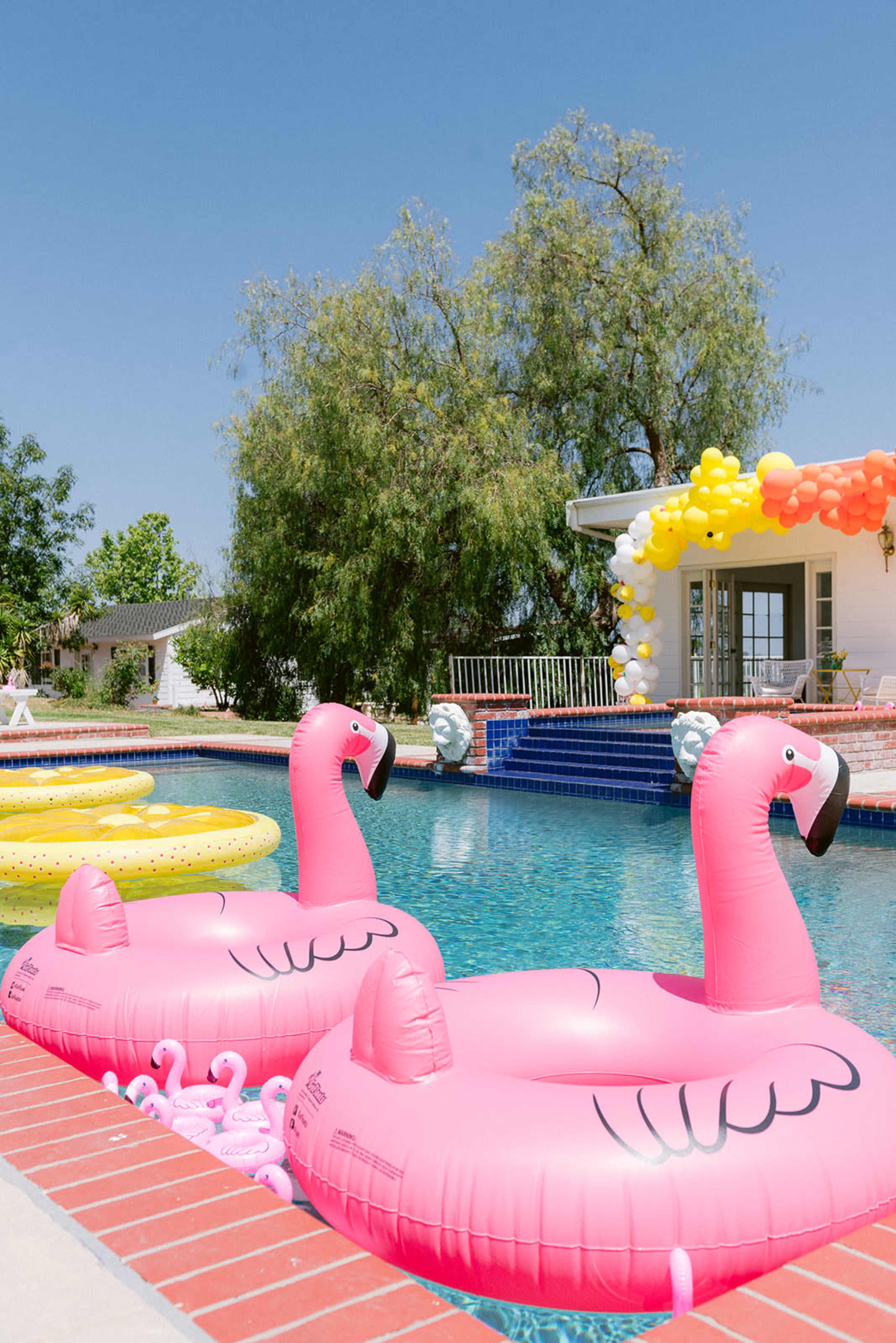 Two pink flamingo-shaped pool floats are positioned in a swimming pool beside a brick edge, with colorful balloons decorating an area in the background.