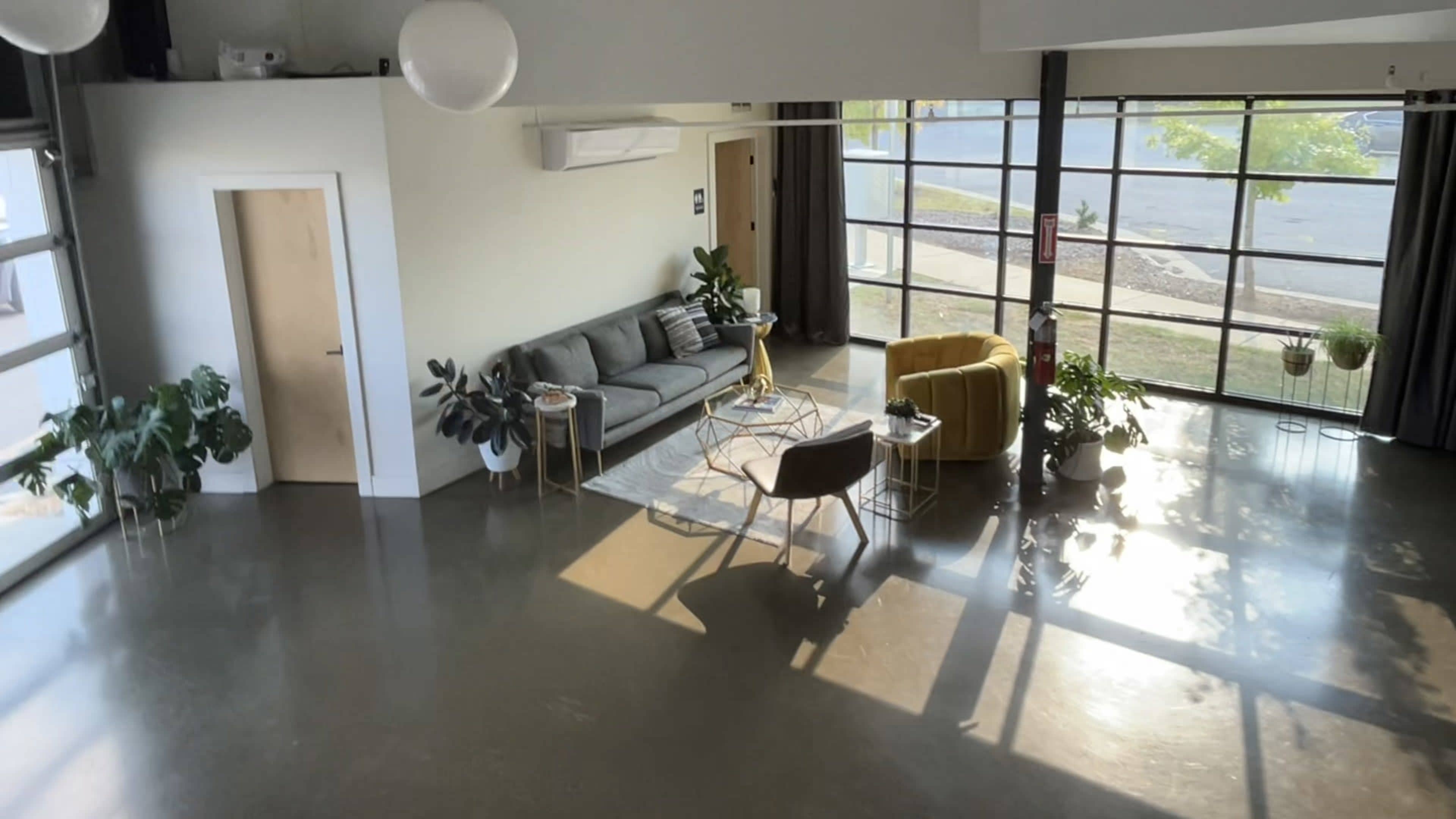A modern living space features a gray sofa and a yellow chair, surrounded by plants and large windows.