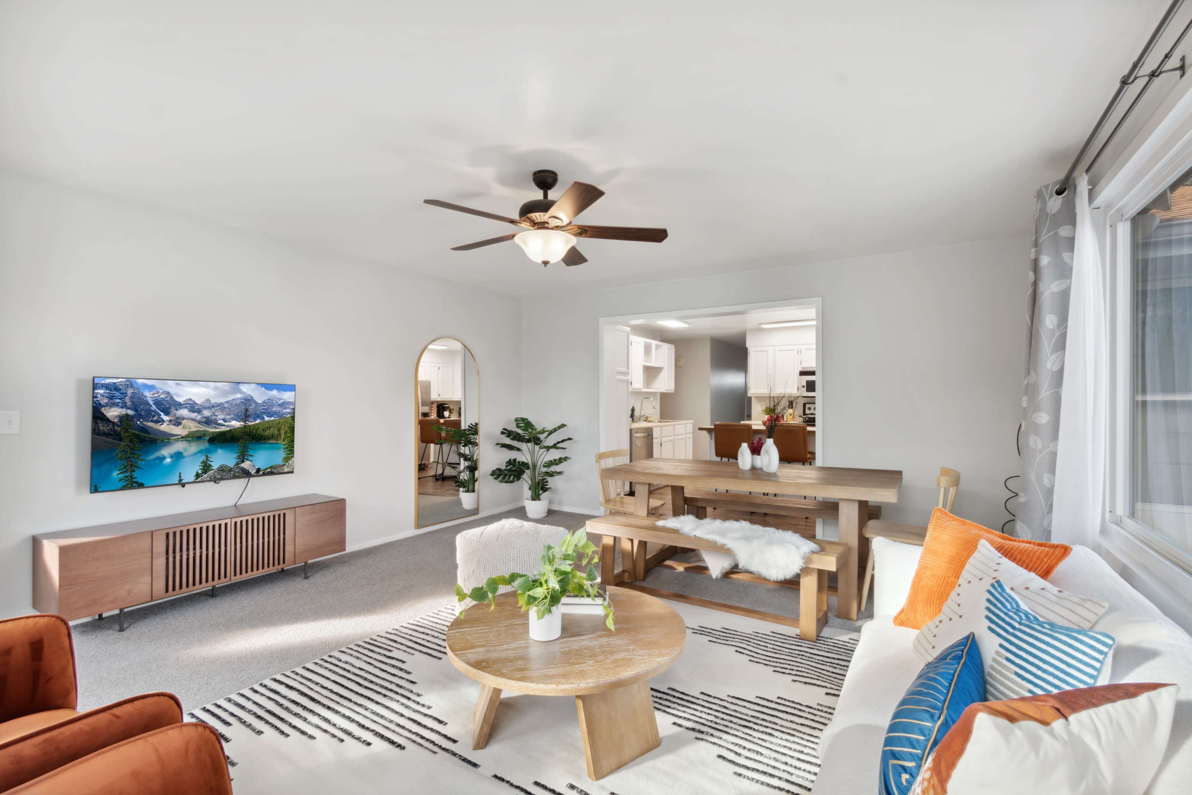 The living room features a television mounted on a white wall, a circular wooden coffee table, a dining table with benches, and an arrangement of furniture centered around a patterned rug.