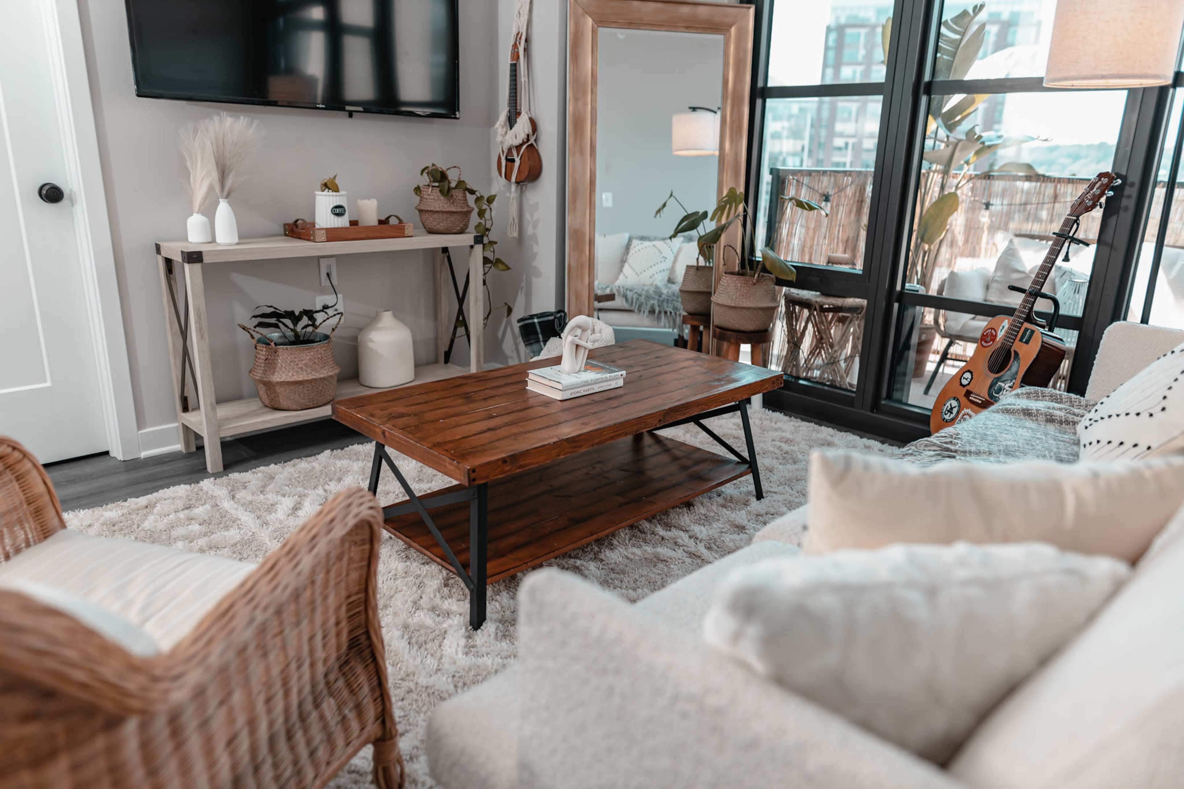 A modern living room featuring a wooden coffee table, a comfortable sofa with decorative pillows, a wall-mounted television, and a large mirror reflecting plants and natural light.