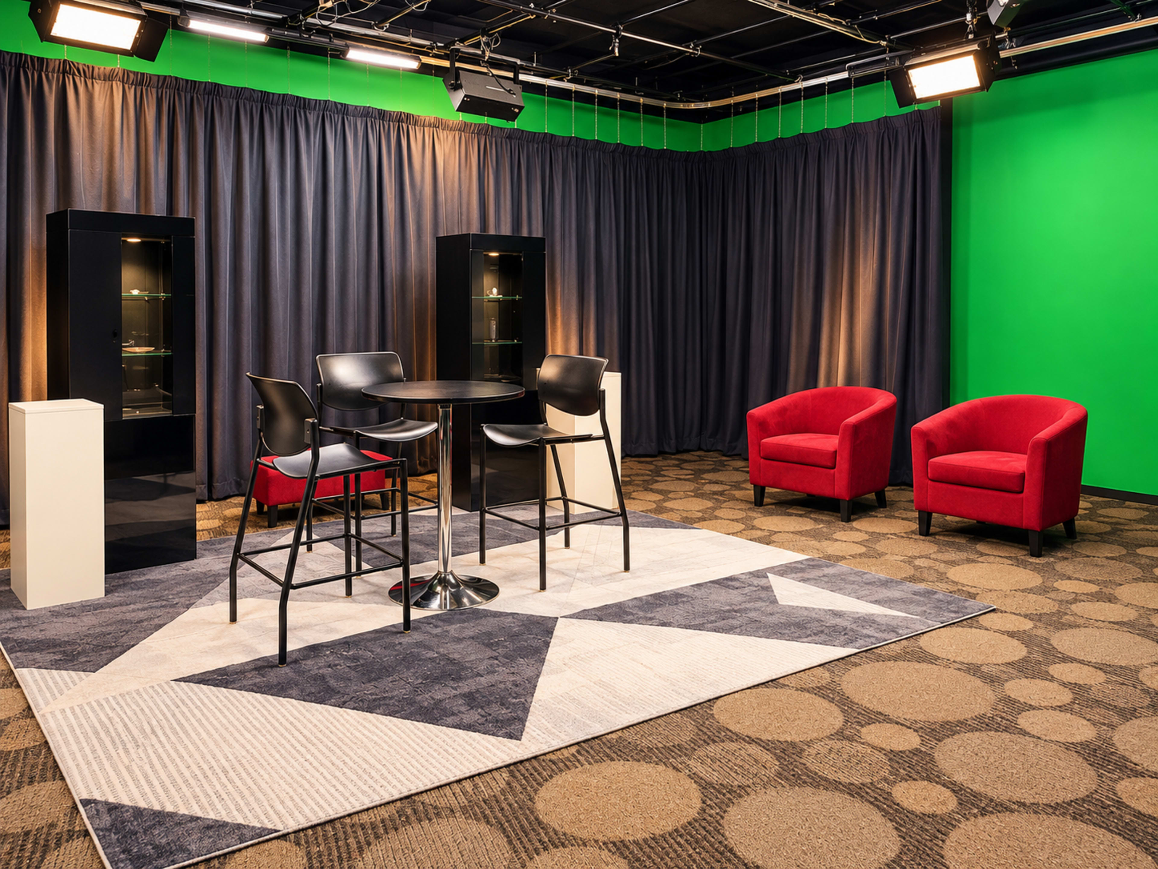 The image shows a studio setup featuring a circular table with black chairs, a patterned rug, two red armchairs, and a green screen backdrop.