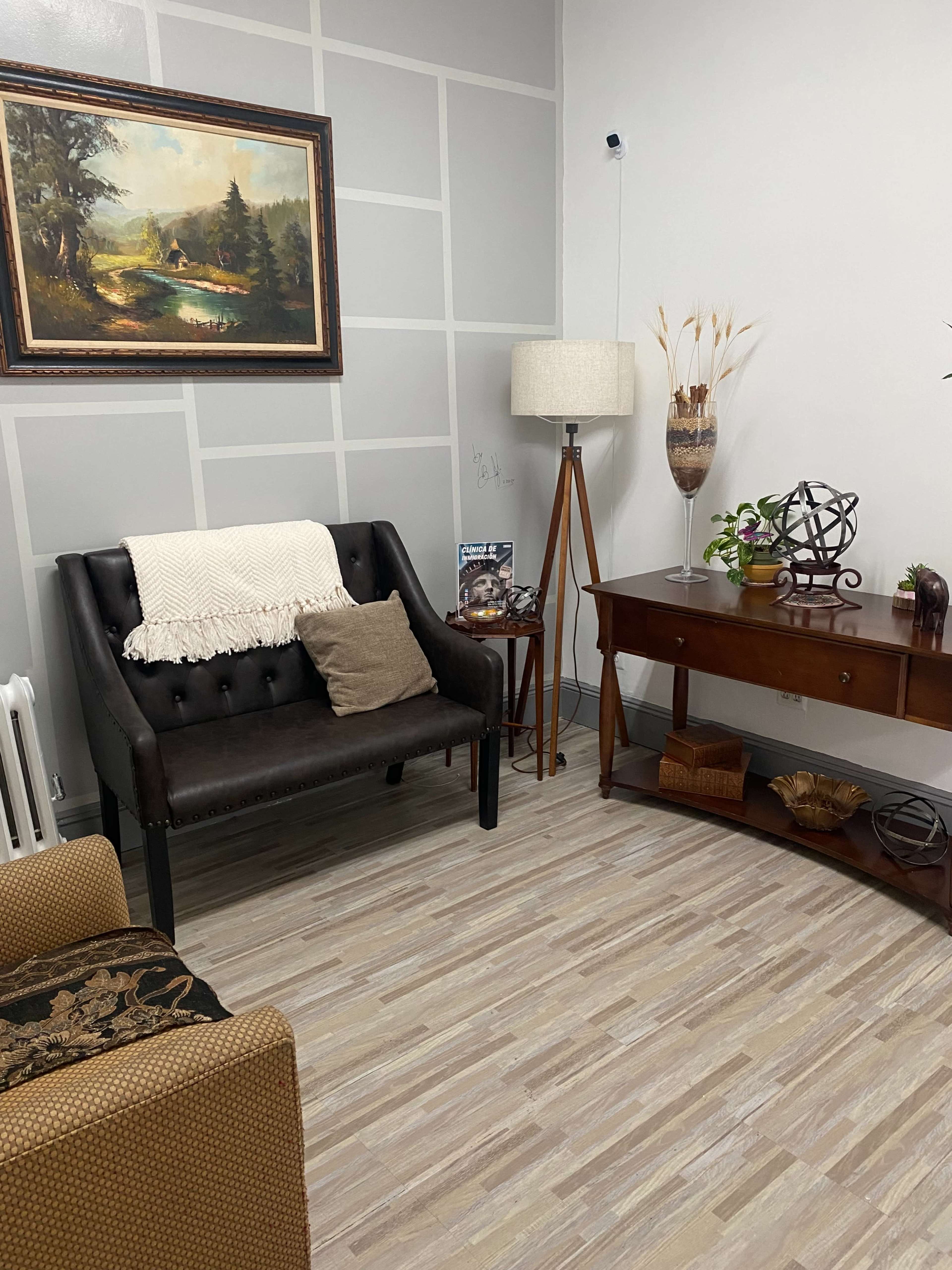 A small, neatly arranged room featuring a black loveseat, a wooden side table, a floor lamp, and a decorative painting on the wall.