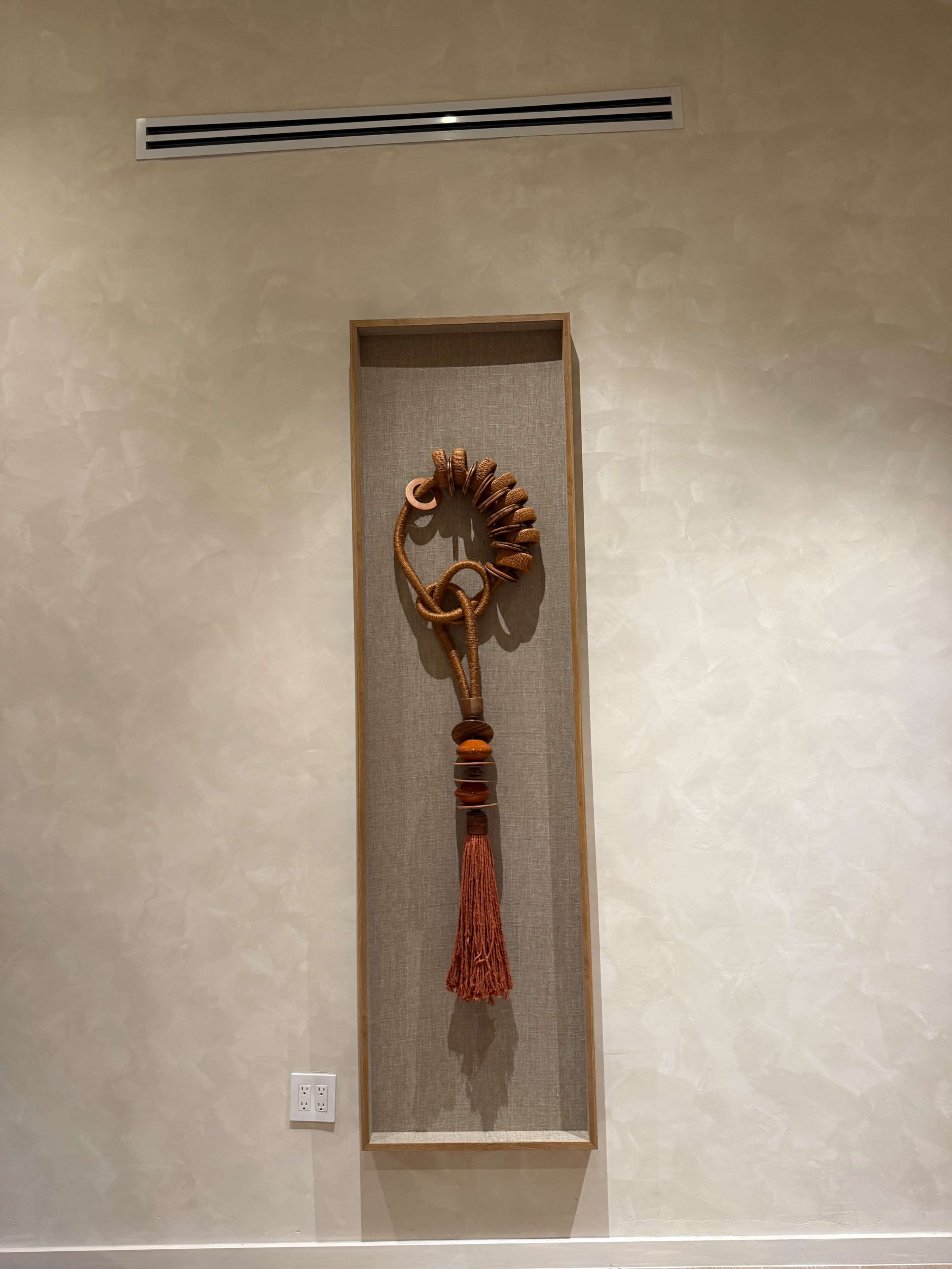 A wooden wall art piece featuring intricate designs and a tassel hangs in a framed alcove on a textured wall.