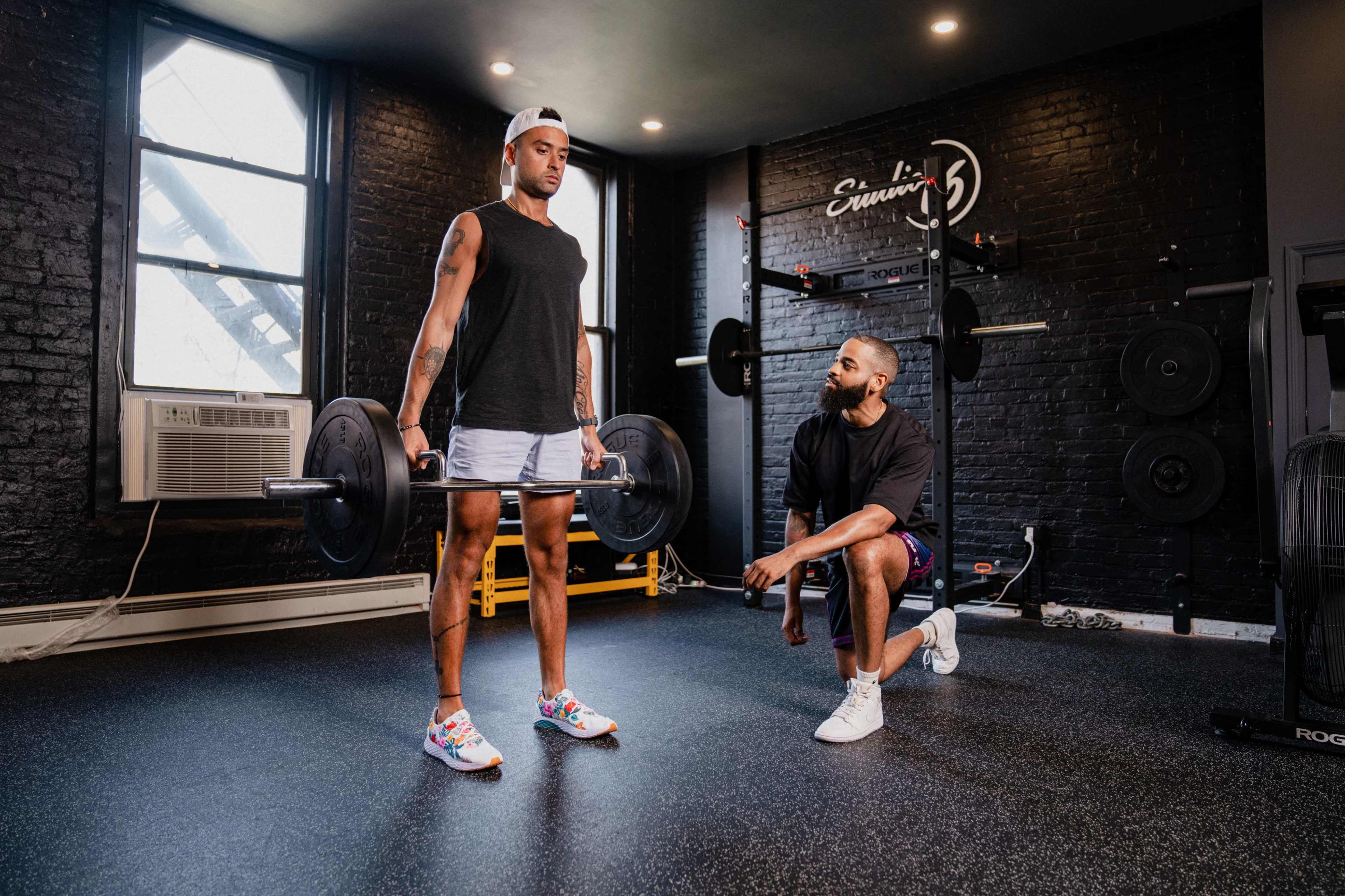 A man performs a deadlift with a barbell while another man kneels beside him, observing in a gym with black walls and equipment in the background.
