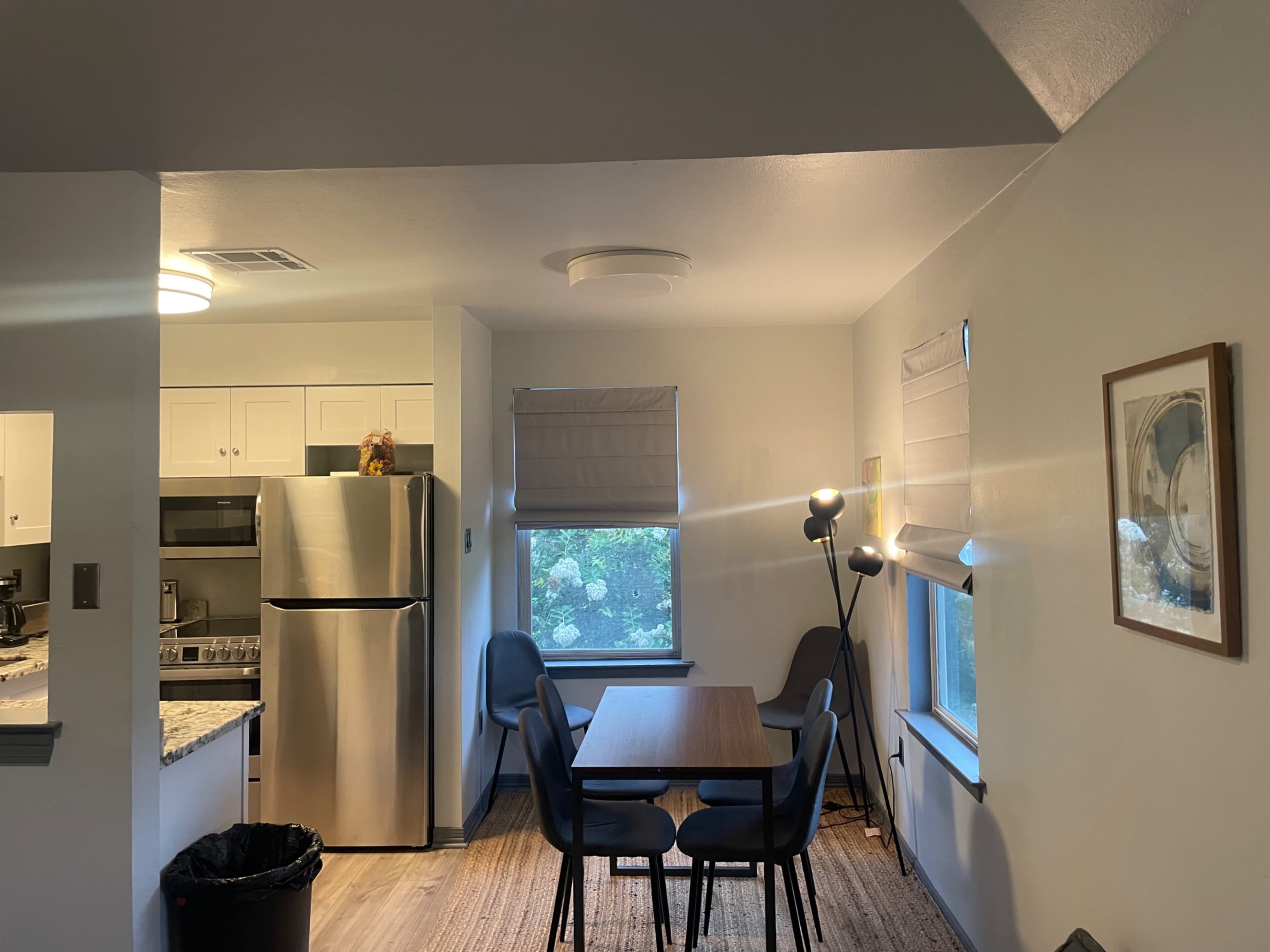 The image shows a dining area with a table and chairs, adjacent to a kitchen featuring stainless steel appliances and a window.