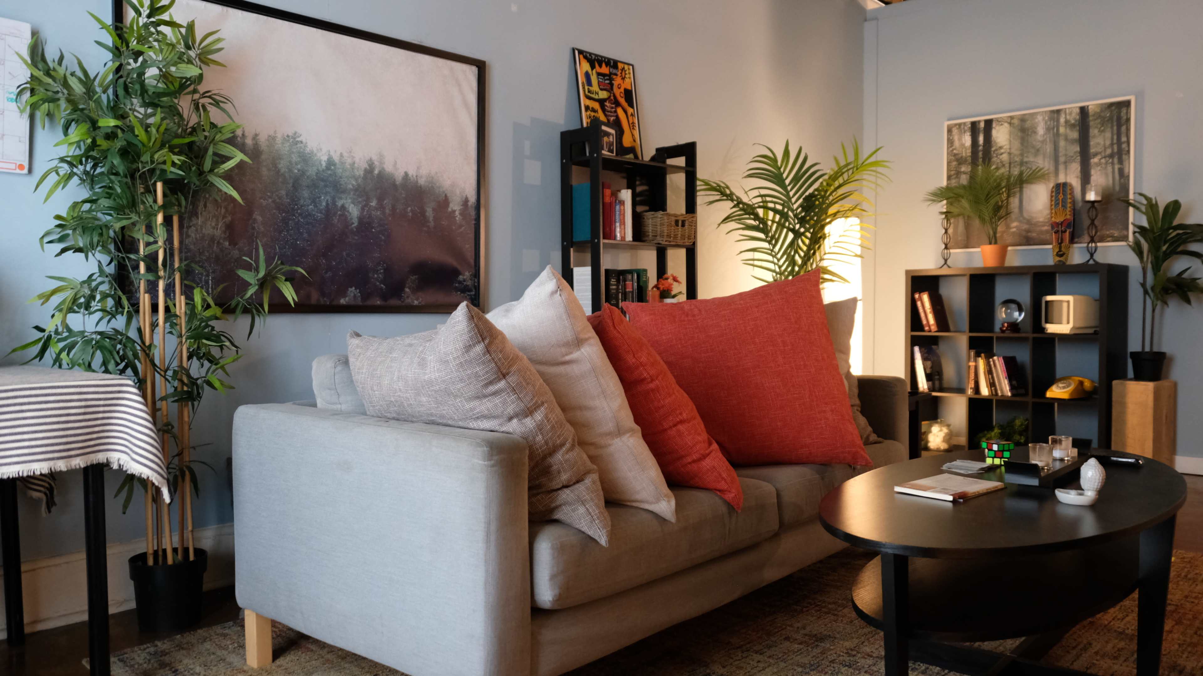 A modern living room features a light gray sofa with large pillows, a round coffee table, and various plants, with bookshelves and artwork on the walls.