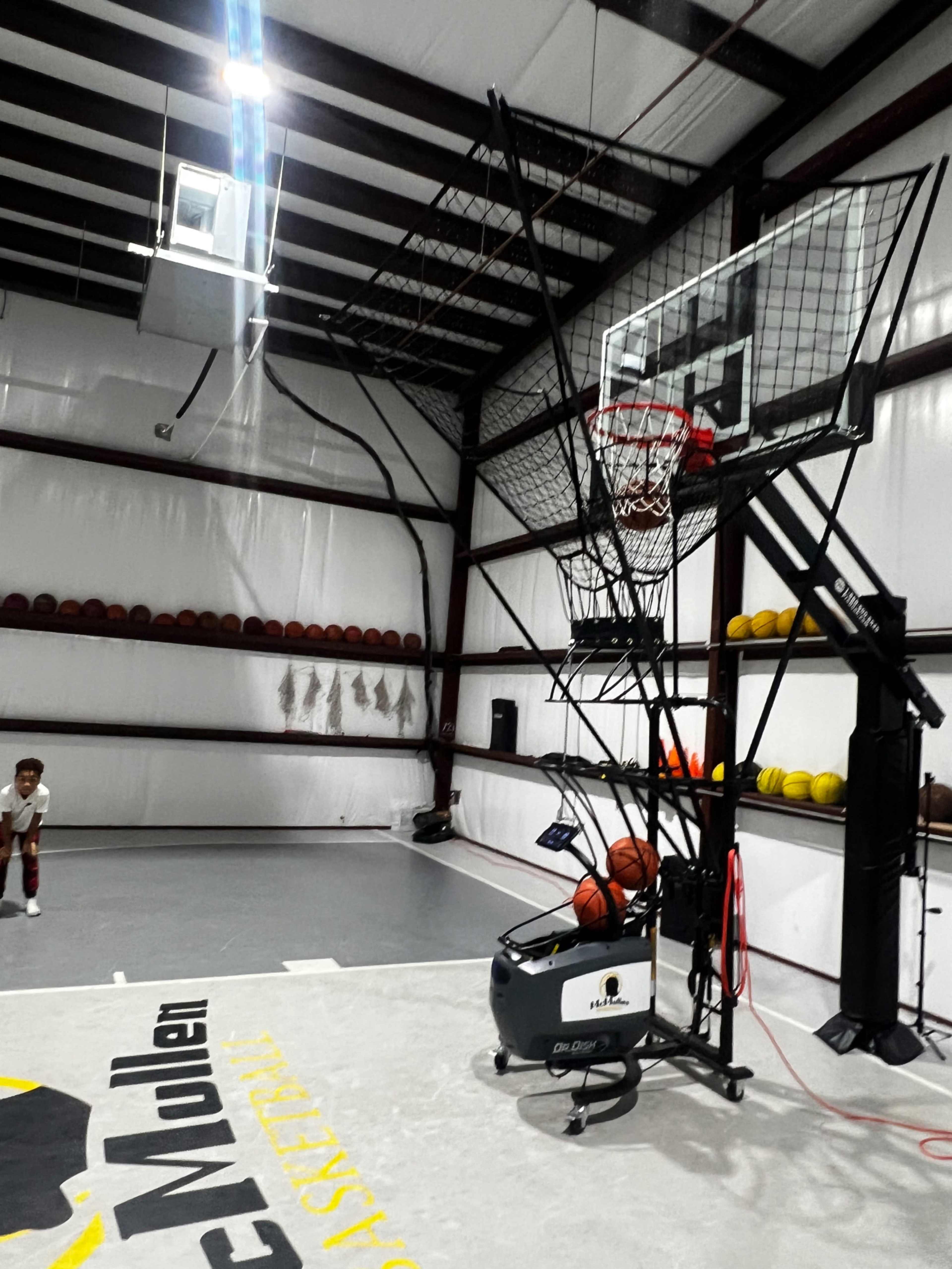 An indoor basketball court features a shooting machine, basketball hoop, and multiple basketballs lined up along the wall.