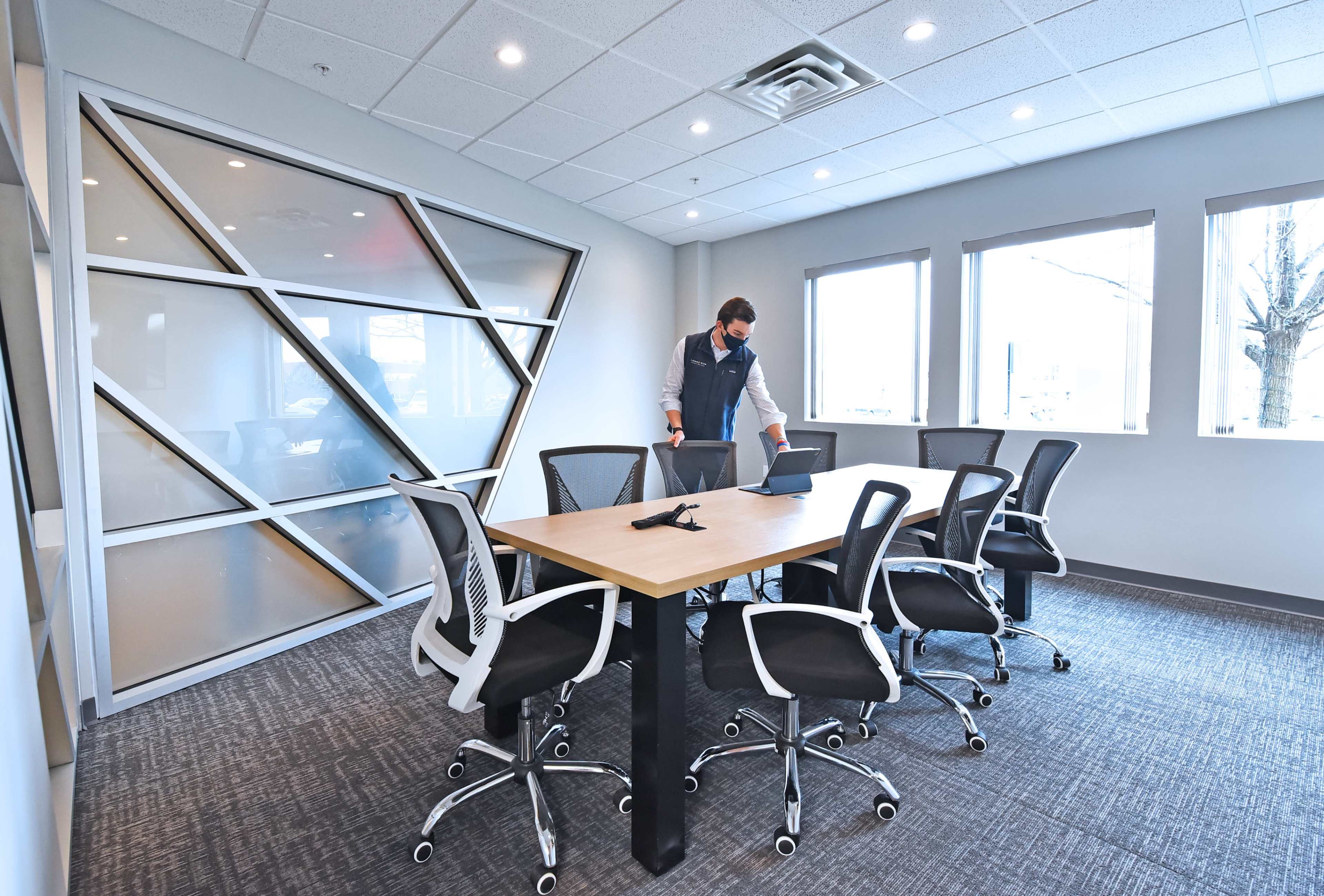 A person is preparing a conference room with a large table and several ergonomic chairs, while standing near a laptop.