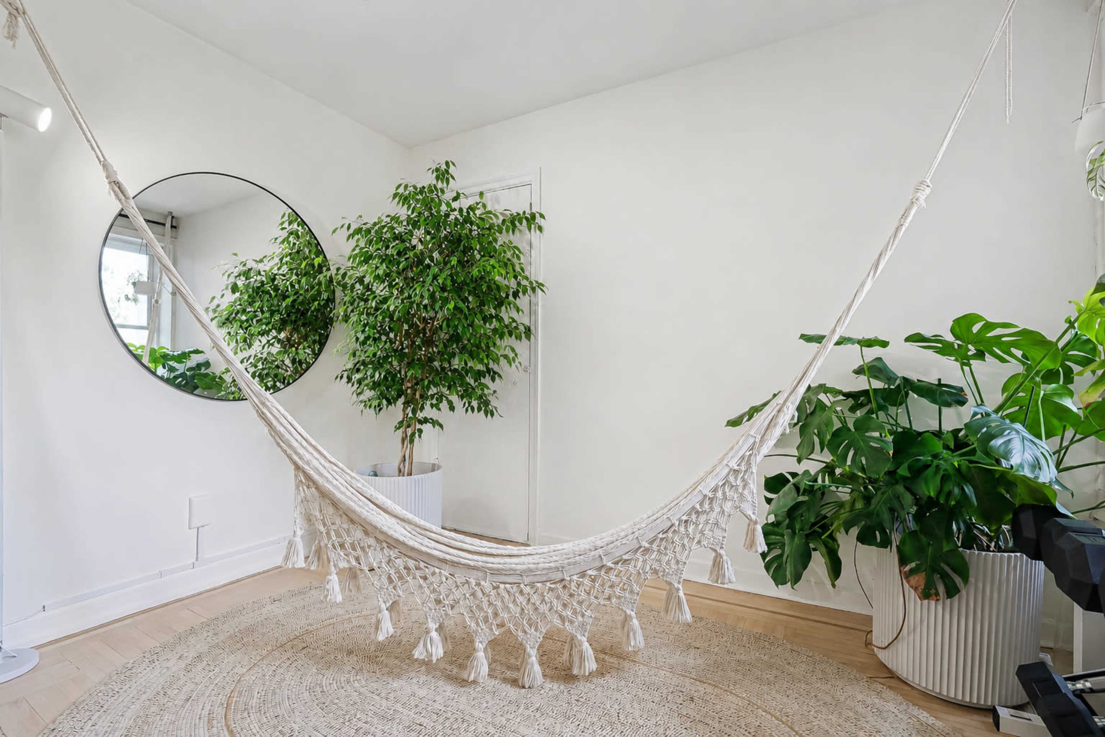 A macramé hammock hangs in a minimalist room adorned with a large mirror and potted plants.