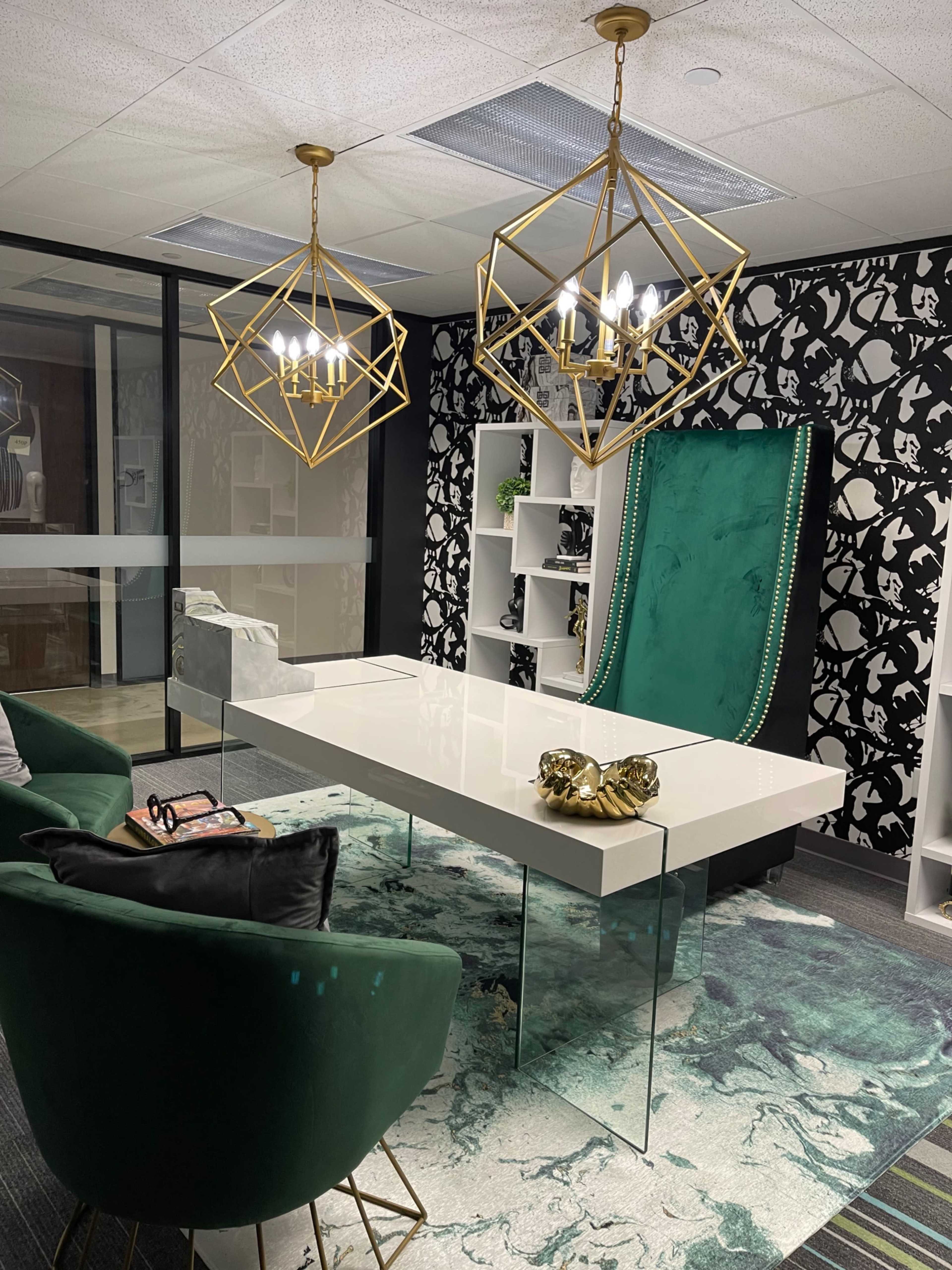 A modern office with a glass-top desk, green velvet chairs, and geometric chandeliers against a patterned wallpaper backdrop.