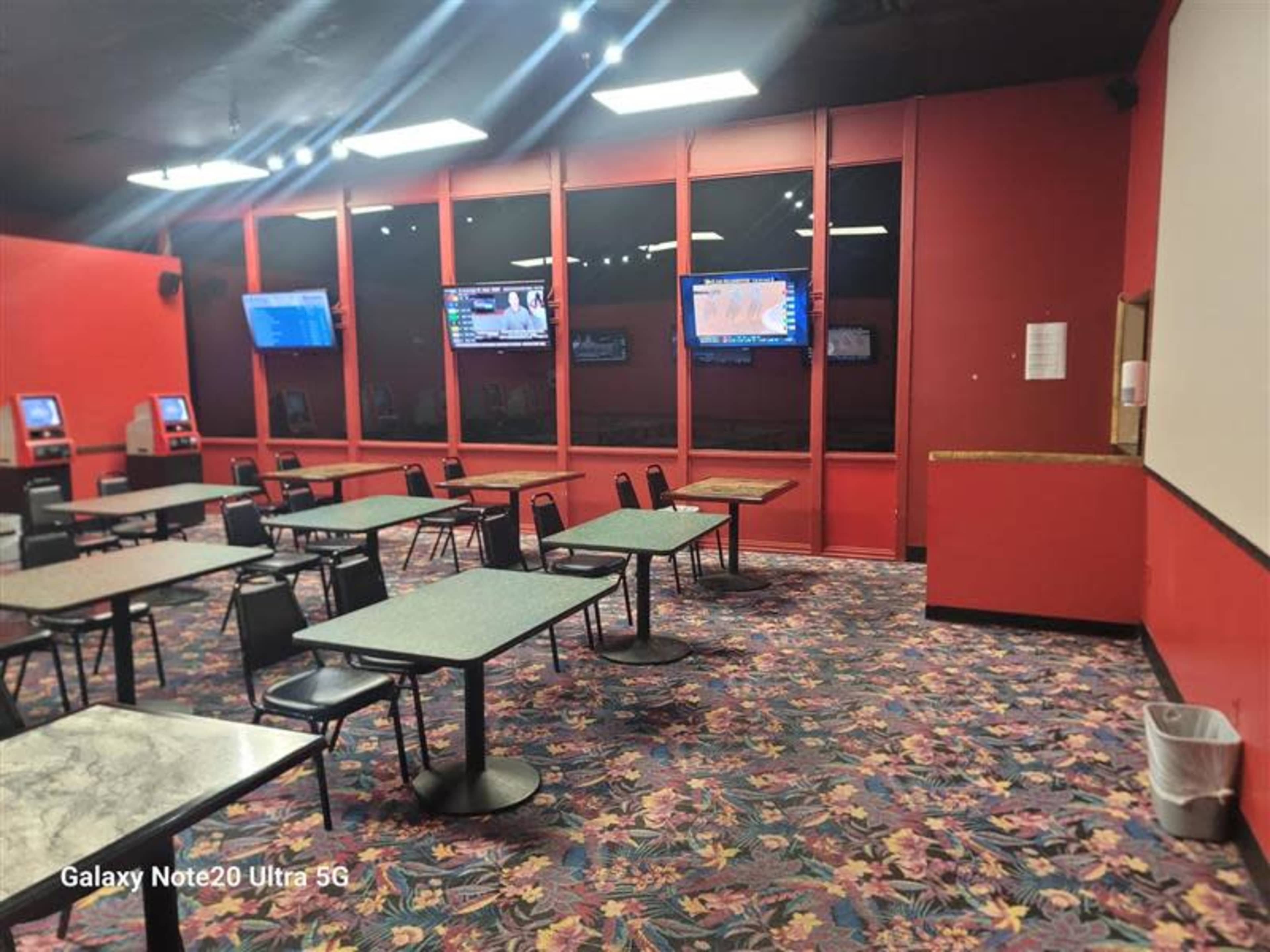 The image shows a brightly colored room with red walls, several tables and chairs arranged on a floral carpet, and two screens mounted on the wall displaying information.