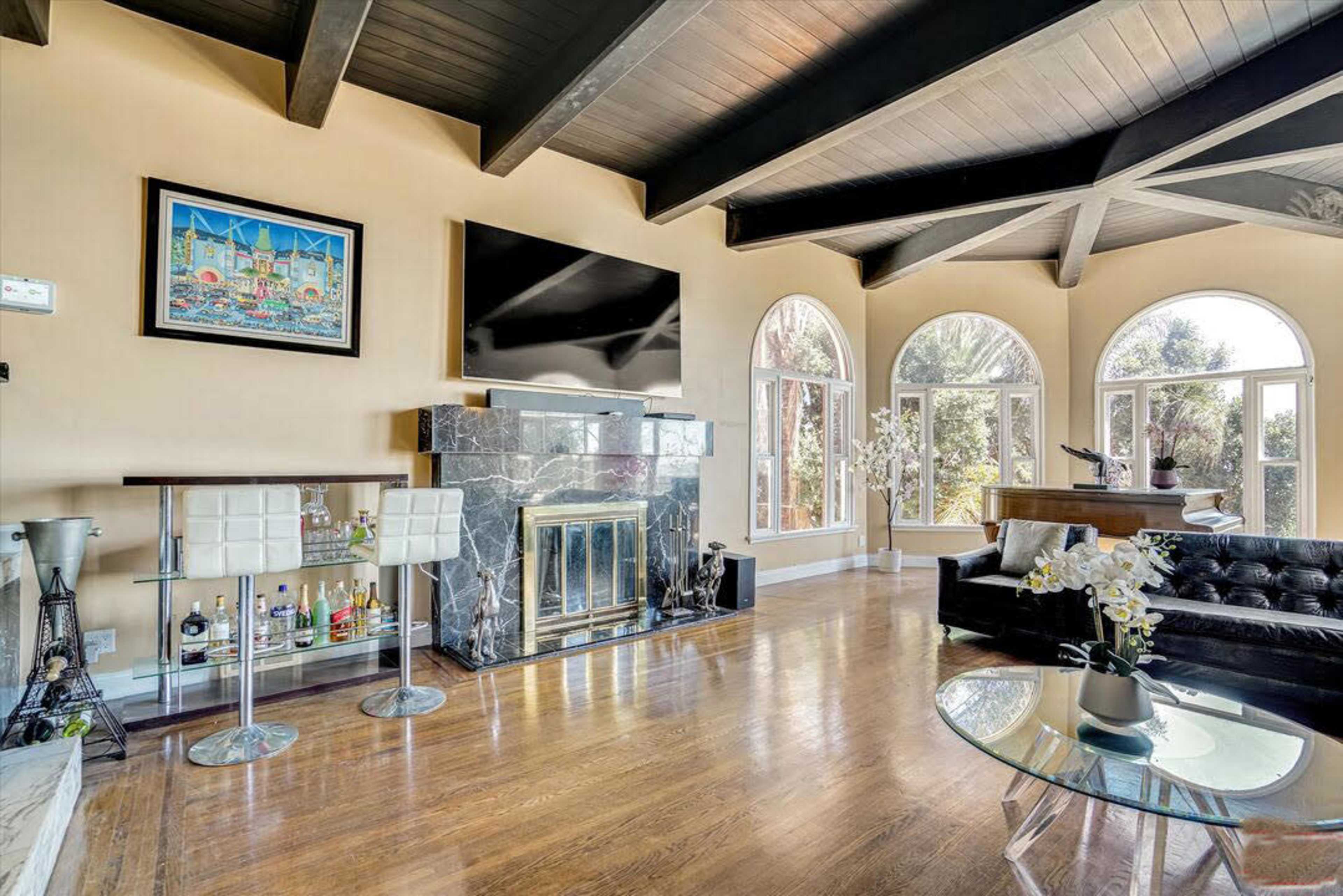 The image shows a living room with a black leather couch, a marble fireplace, a large TV mounted on the wall, and arched windows allowing natural light to enter.