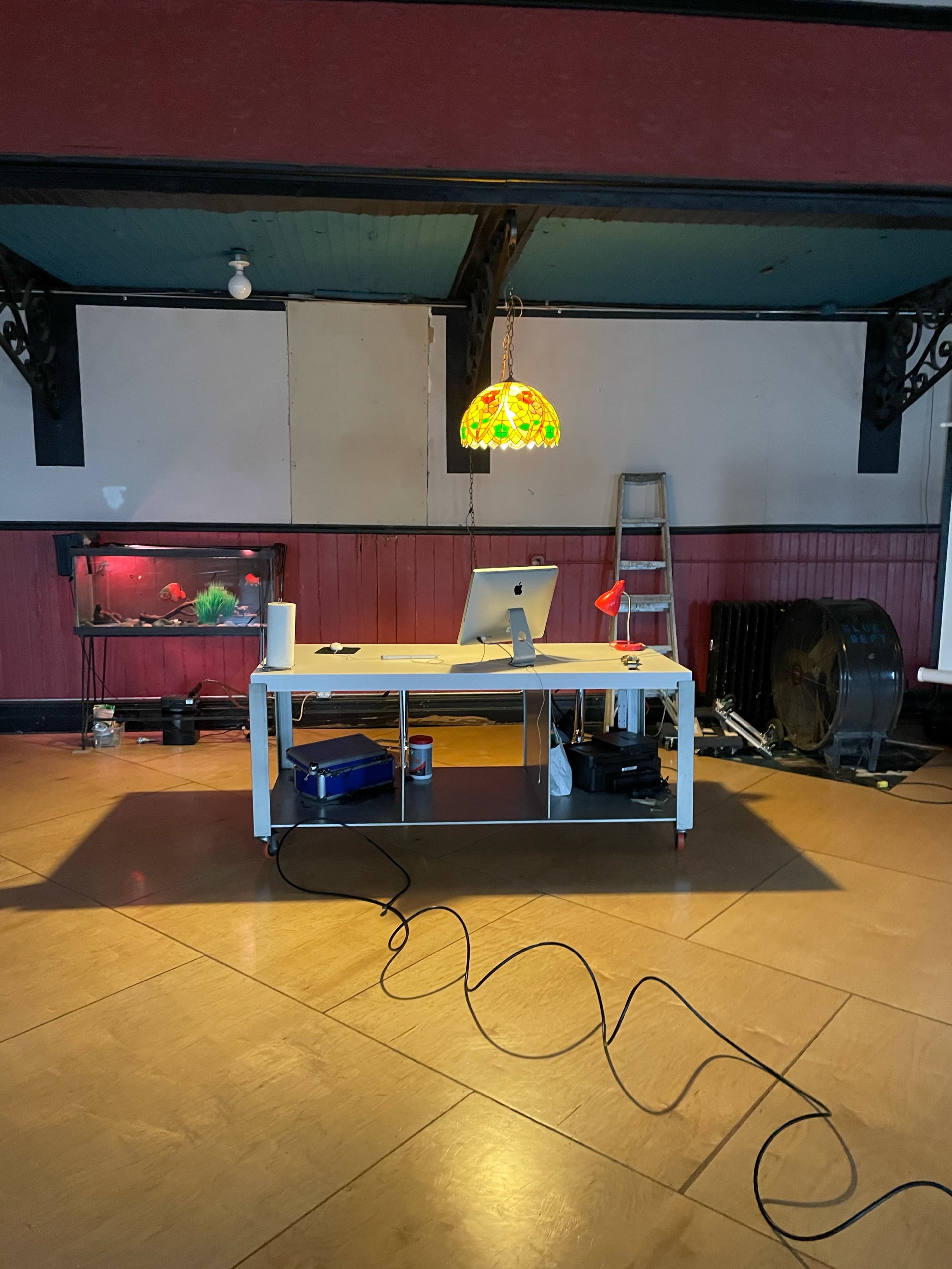 A workspace featuring a desk with a computer, a colorful lamp, and a fish tank, set against a partially decorated wall and a ladder in the background.