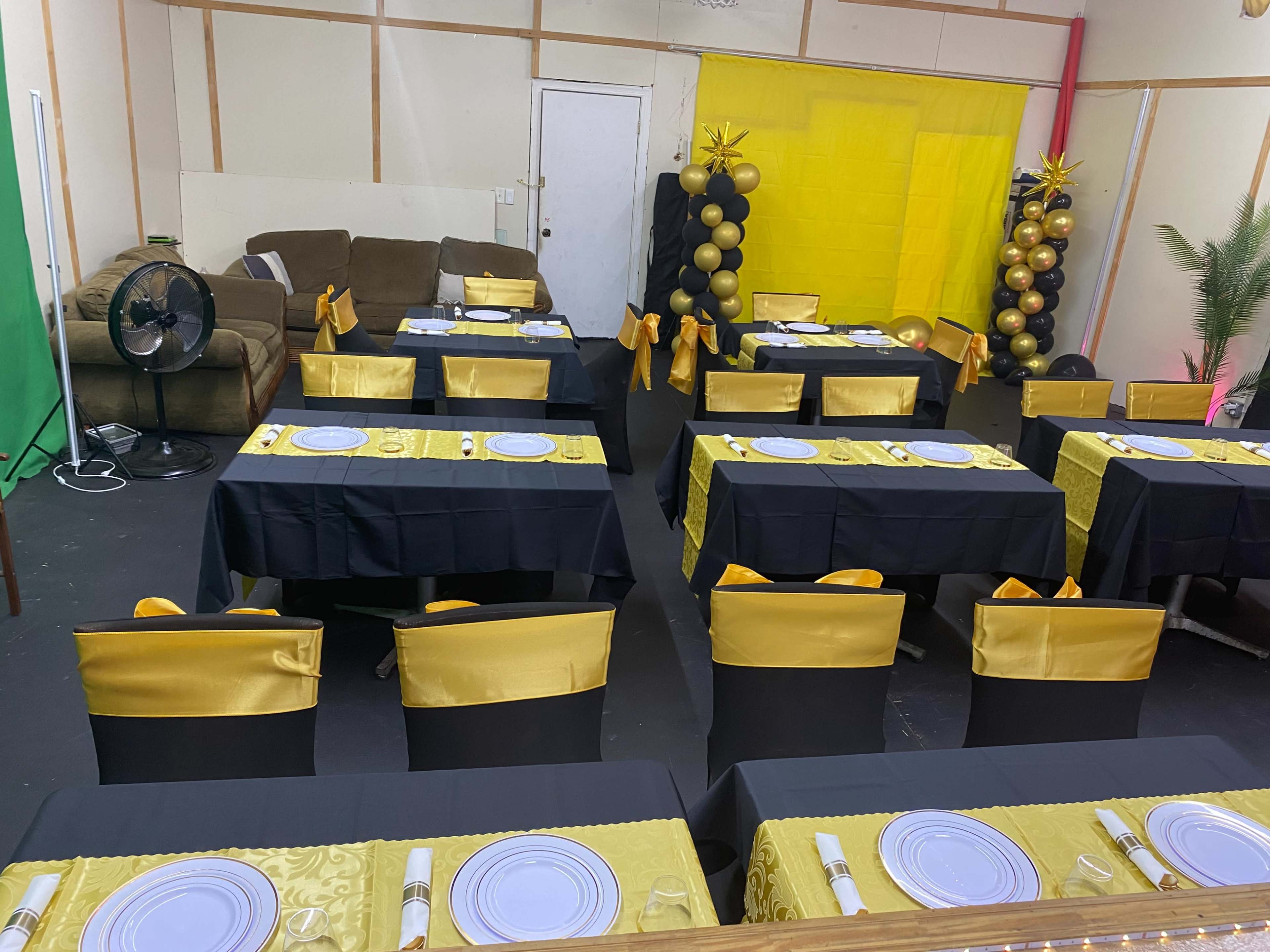 The image displays a decorated event space with several tables set in black and gold, complete with place settings, a yellow backdrop, and a couch in the corner.