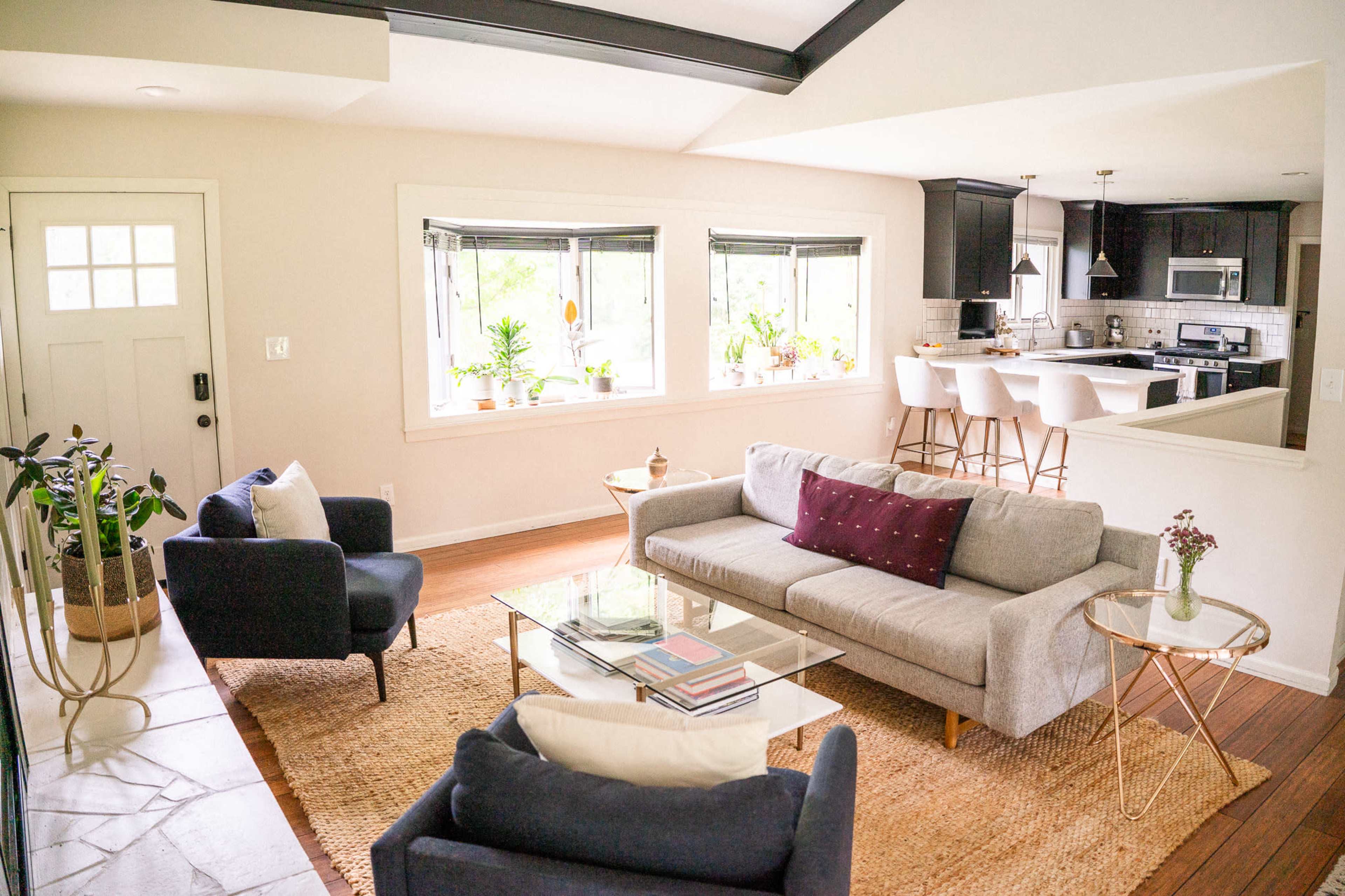 A modern living room with a gray sofa, two dark chairs, a glass coffee table, and an open kitchen visible in the background.