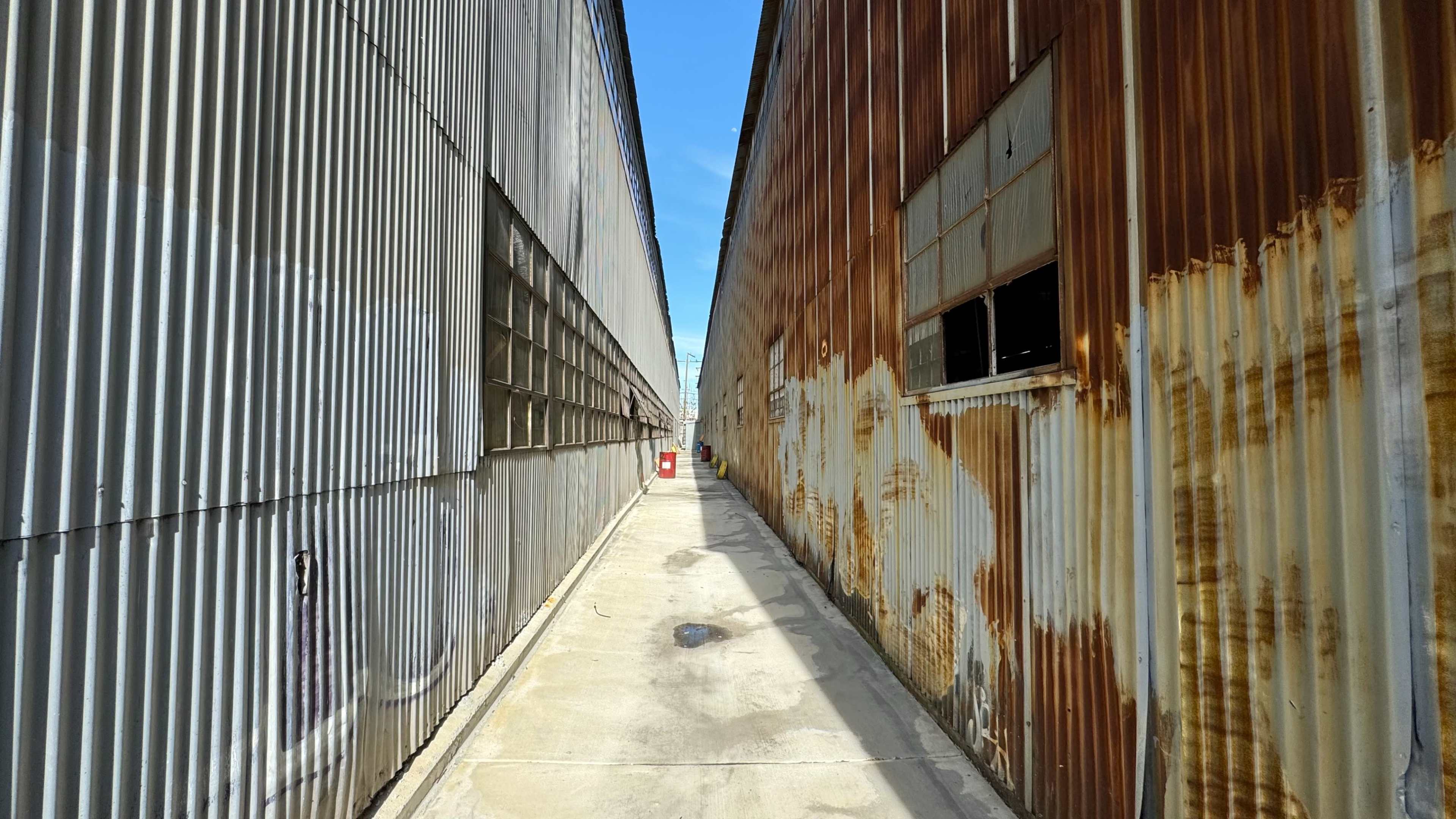 A narrow passageway separates two corrugated metal structures, one weathered and rusty, under a clear blue sky.