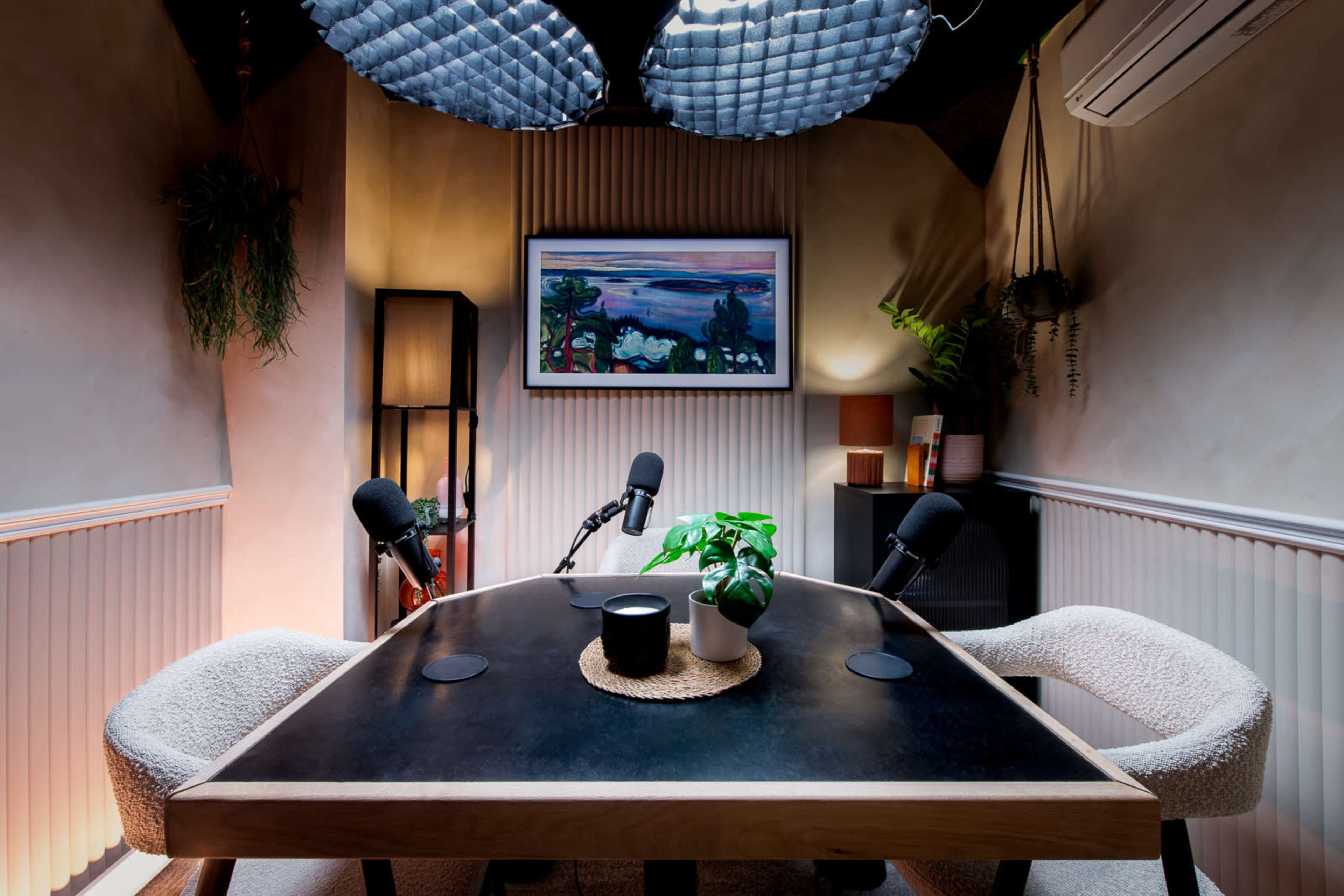 A podcast studio features a wooden table with two chairs, microphones set up, and a framed artwork on the wall, surrounded by plants and ambient lighting.