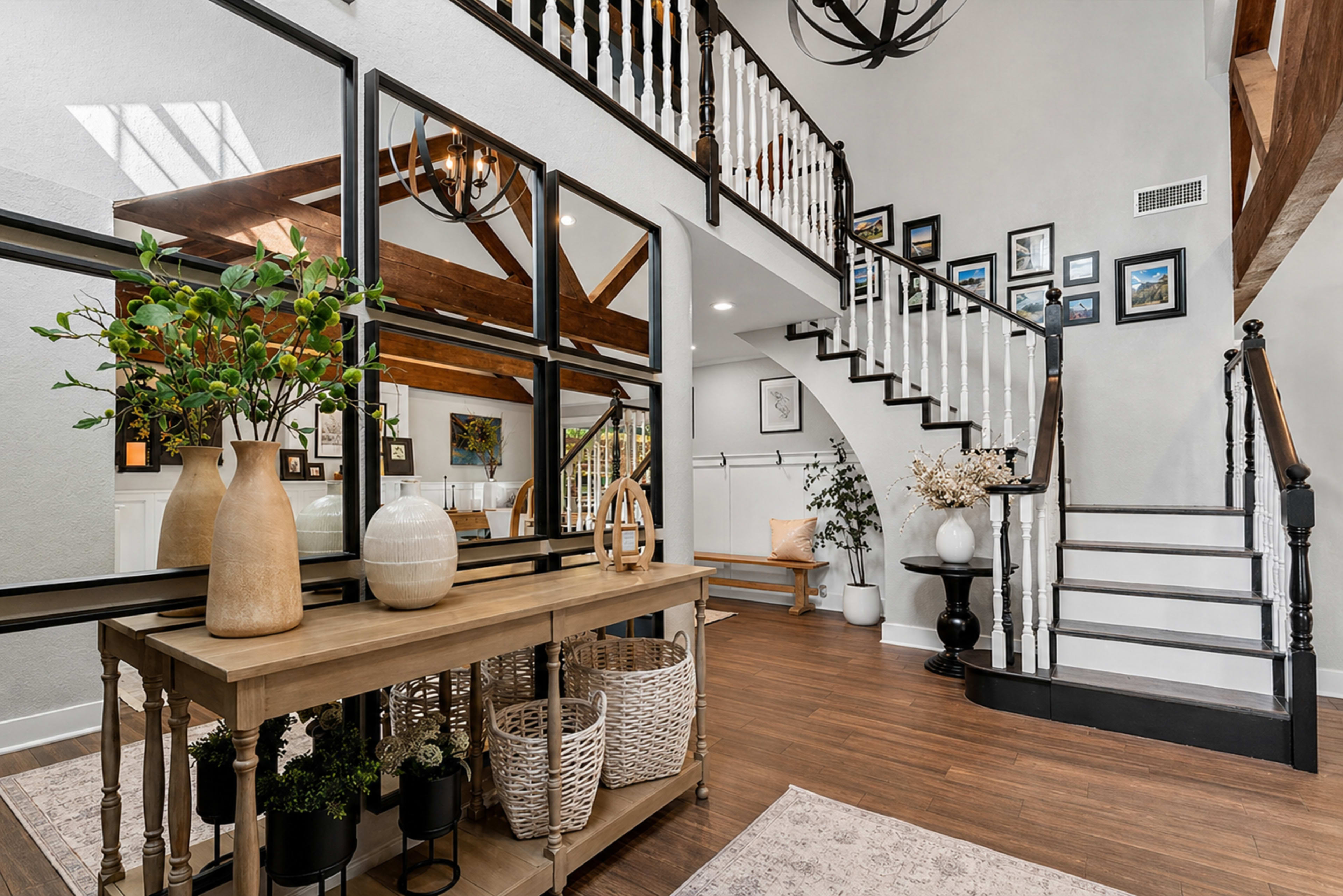 The image shows a spacious entryway featuring a wooden table with decorative vases, a staircase with a black railing, and framed pictures on the wall.