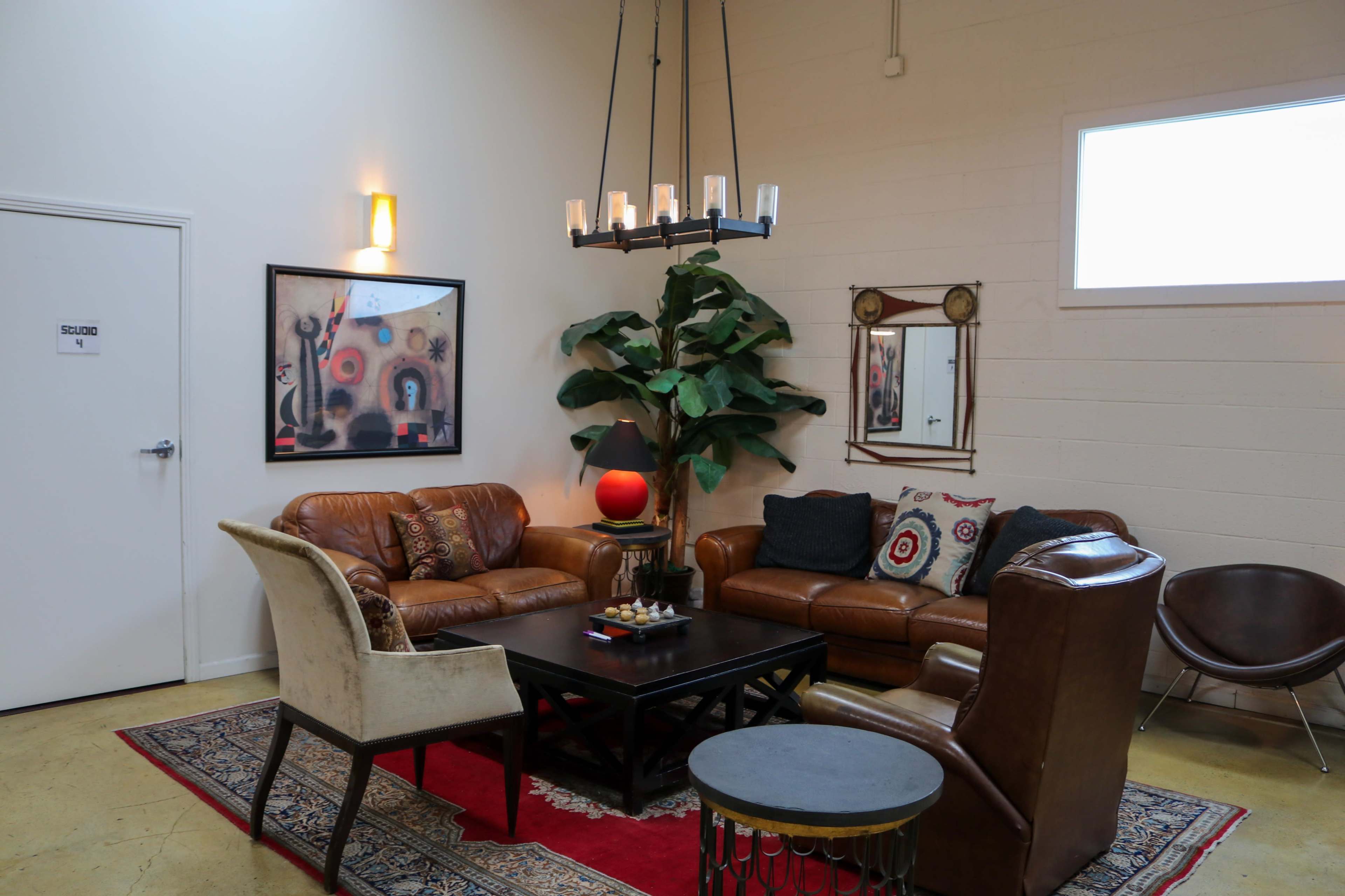The image shows a cozy lounge area featuring leather sofas, a coffee table, and a large plant, complemented by decorative wall art and a chandelier.