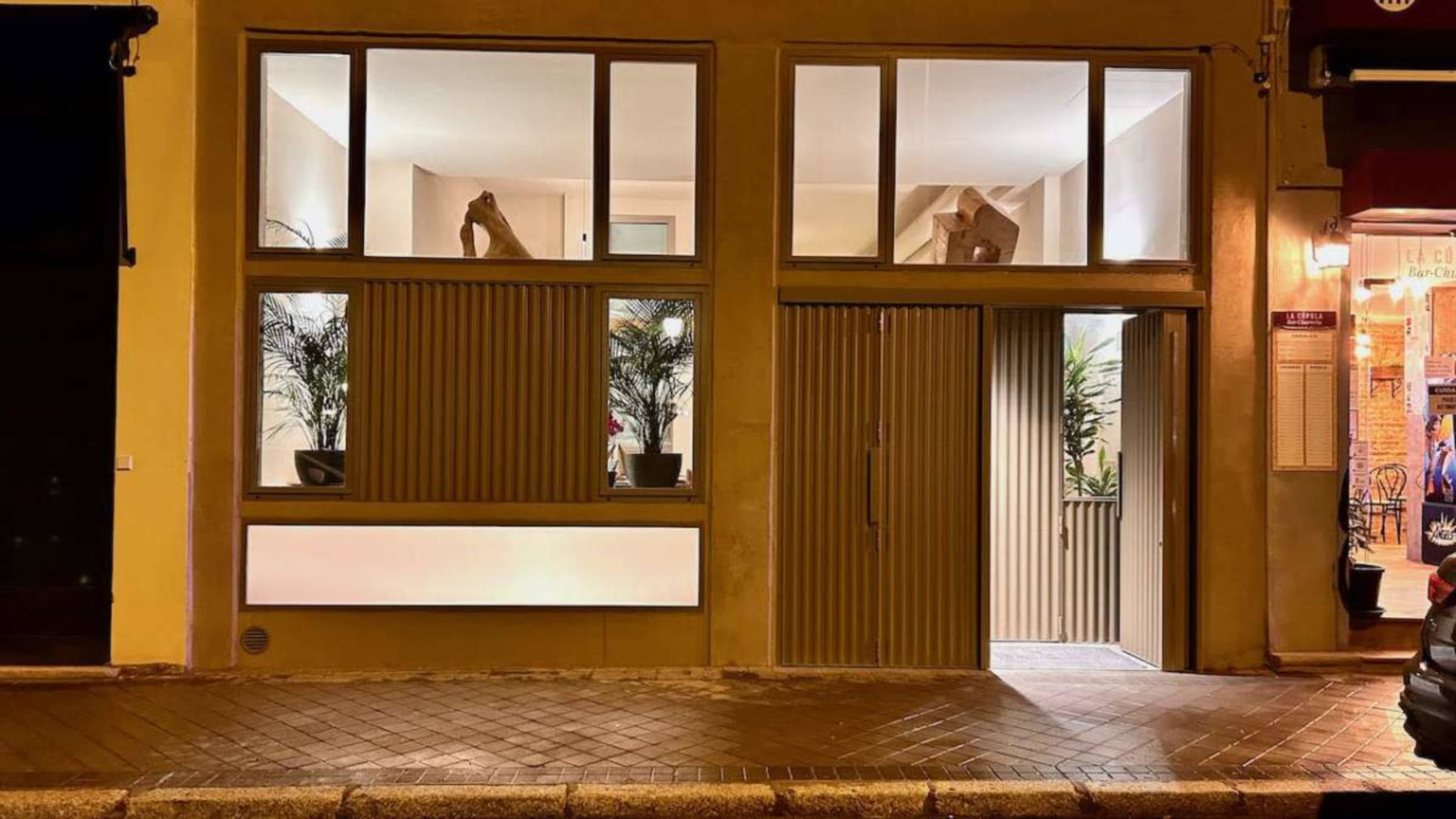 The image shows a modern building facade with large windows, decorative plants, and a closed door on a paved street at night.