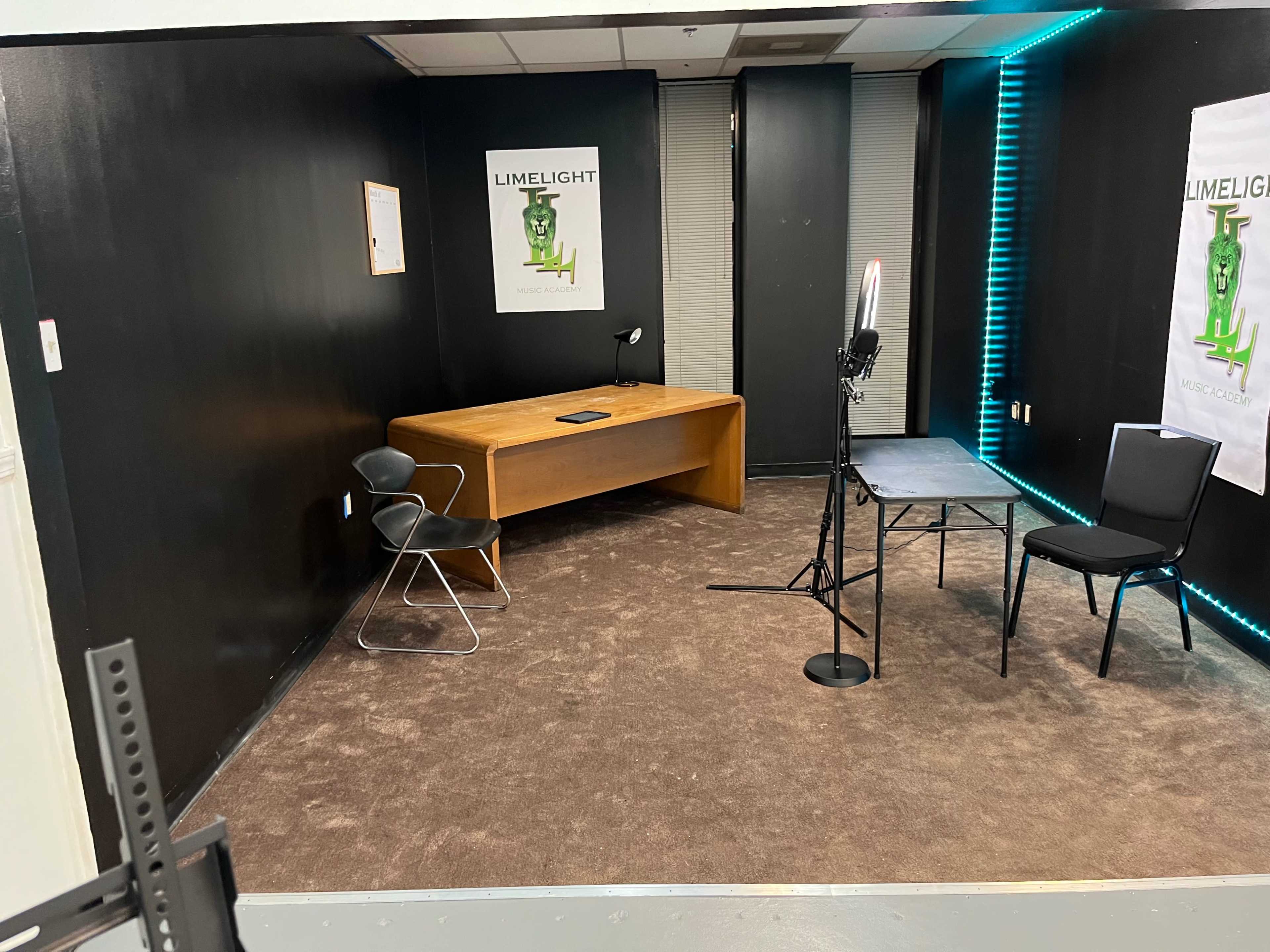 The image shows a small, dimly-lit room featuring a wooden desk, two chairs, and a microphone setup, with posters on the walls.