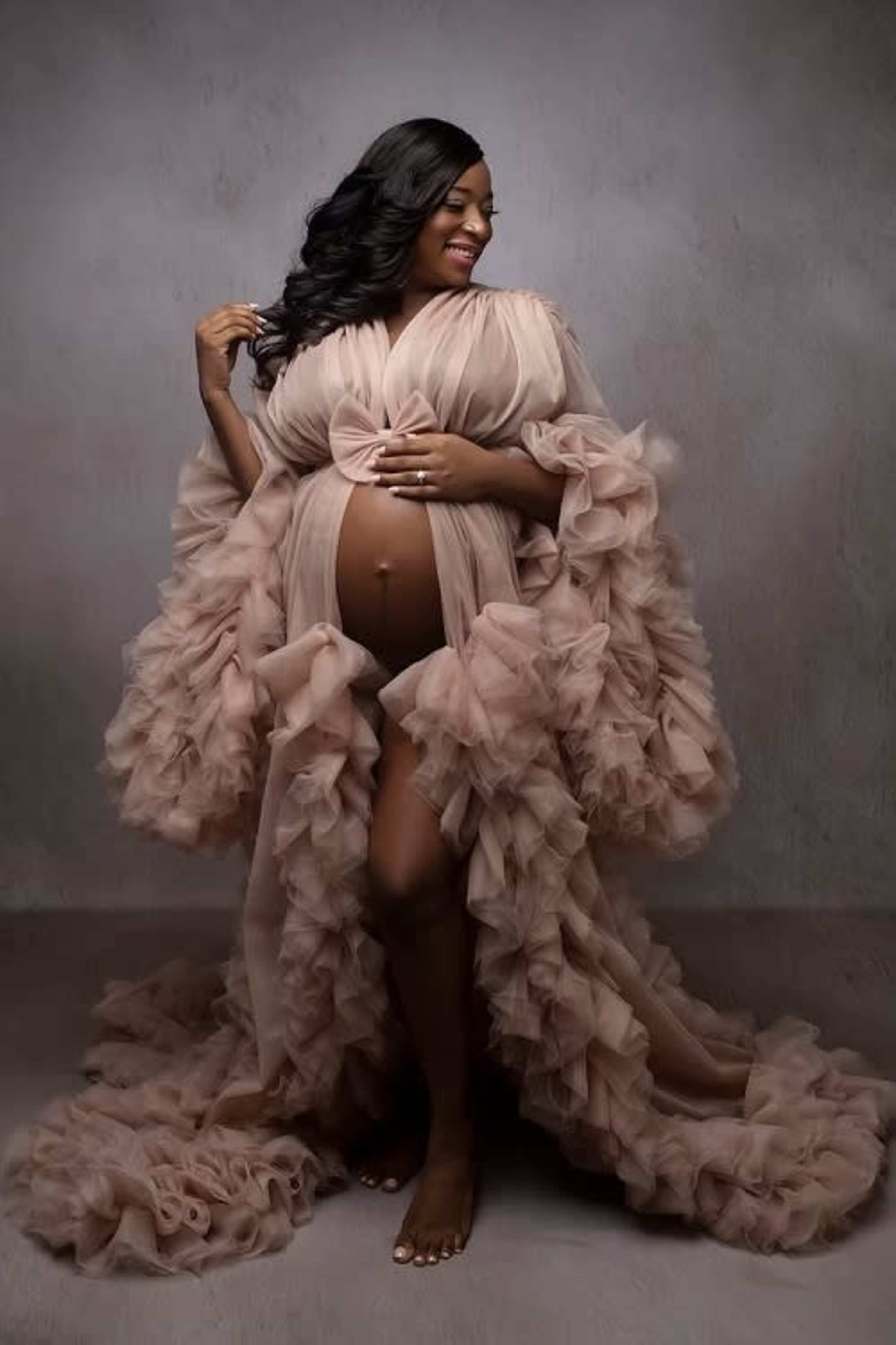 A pregnant woman poses in a flowing, ruffled gown against a gray backdrop.