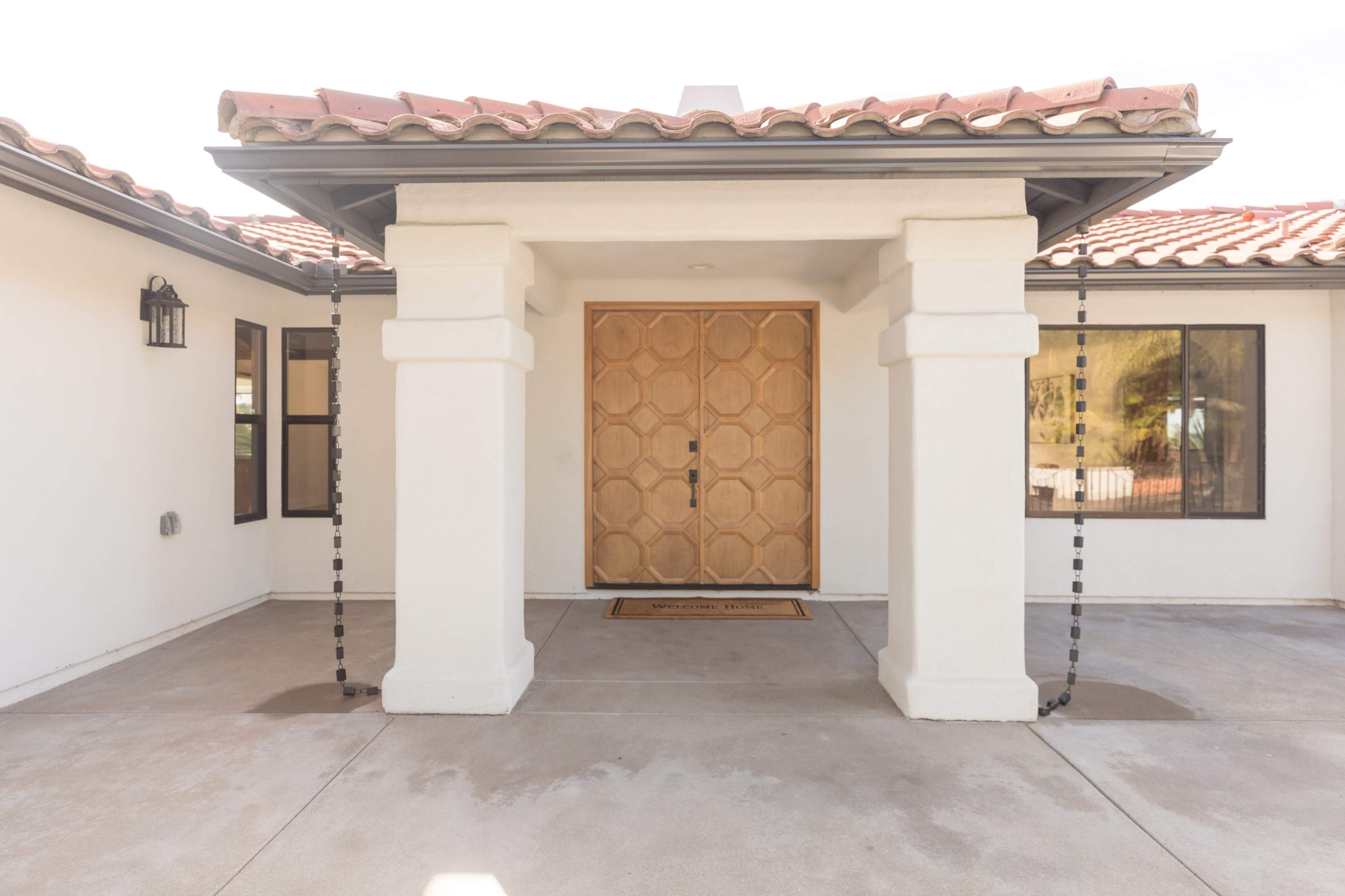 The image shows a modern entrance to a house featuring a large wooden door with geometric patterns, flanked by two windows and a tiled roof.