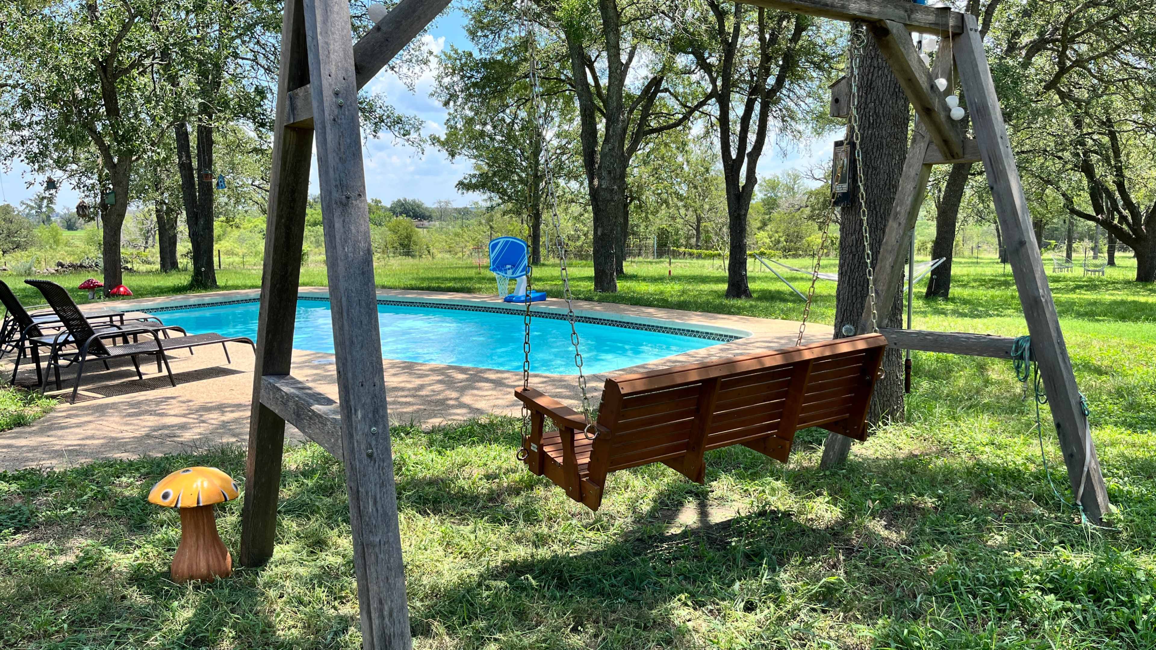 A wooden swing hangs near a swimming pool surrounded by green grass and trees.