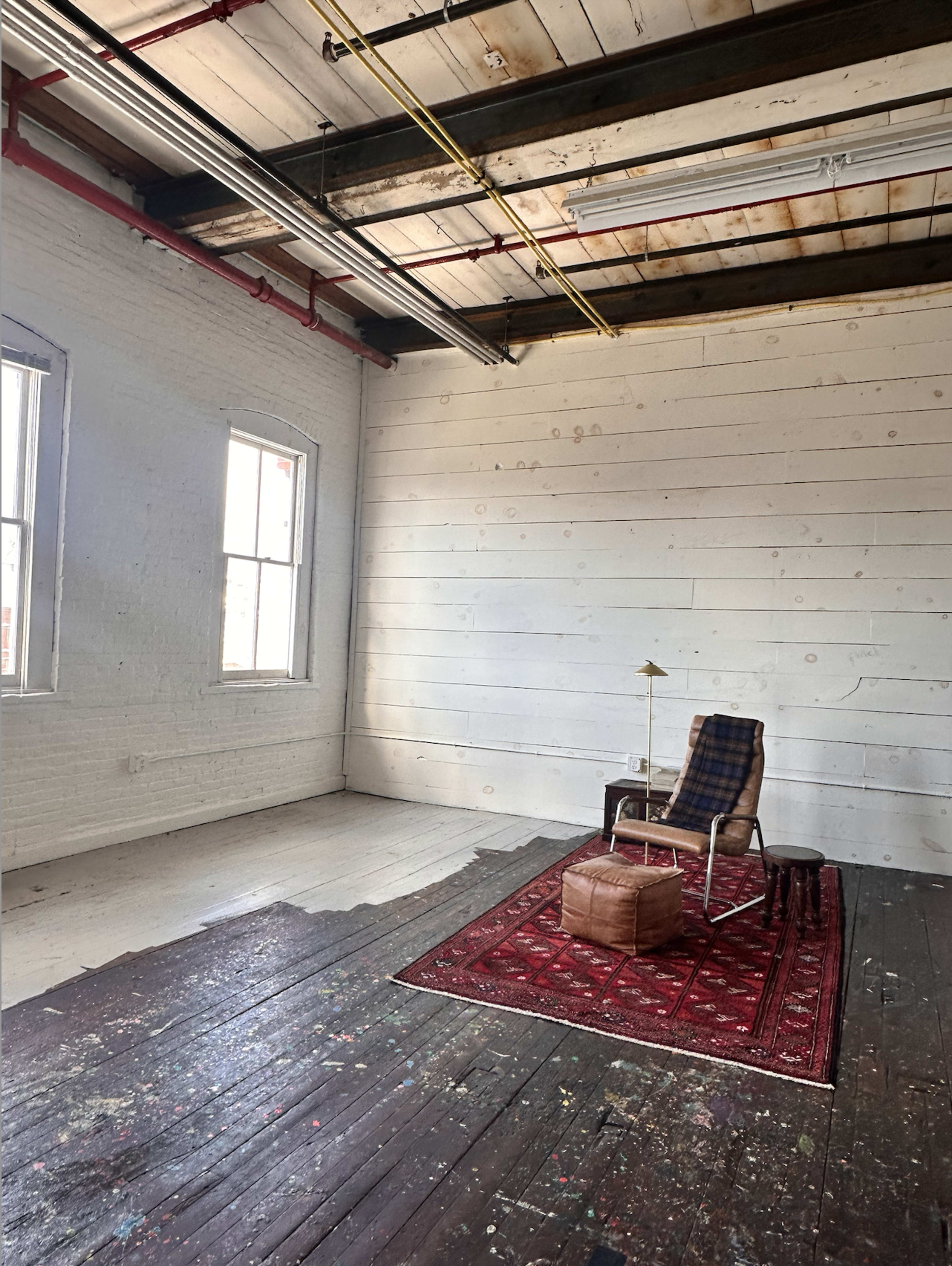 A sparsely furnished room features a wooden floor, two windows, a rug, a chair with a plaid cushion, and a small table lamp.