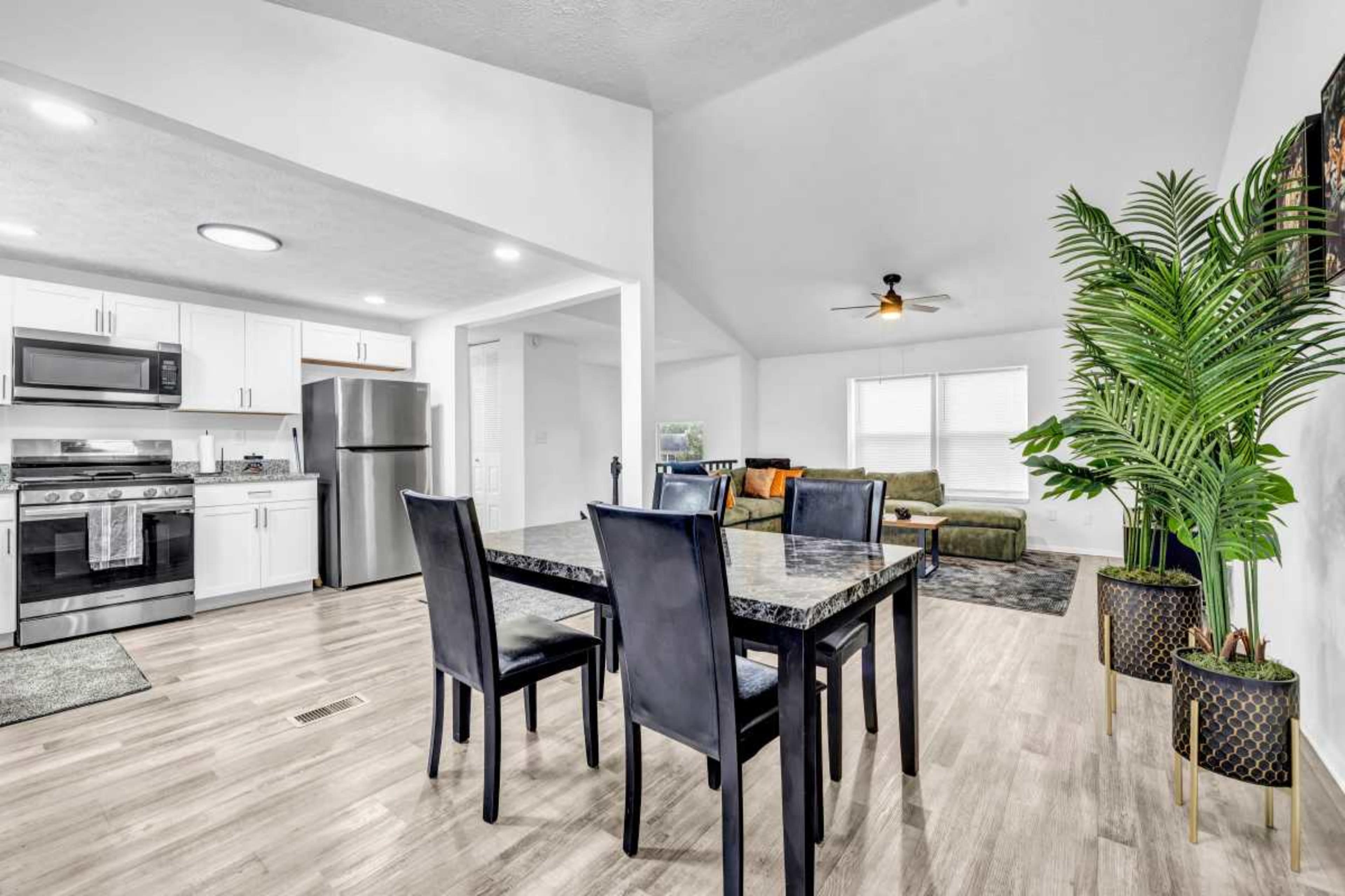 A modern dining area features a dark table and chairs, with a kitchen in the background and a living room with a sofa and greenery in the foreground.