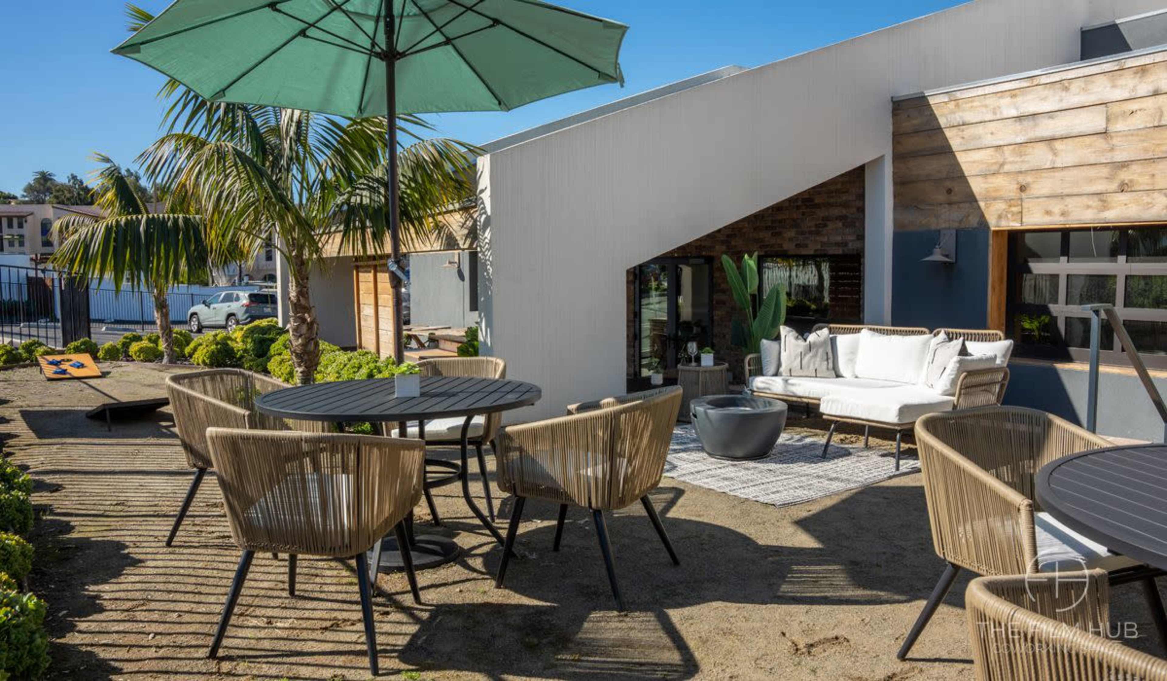 A modern outdoor seating area with round black tables, a large green umbrella, and a comfortable lounge setup beside a palm tree.