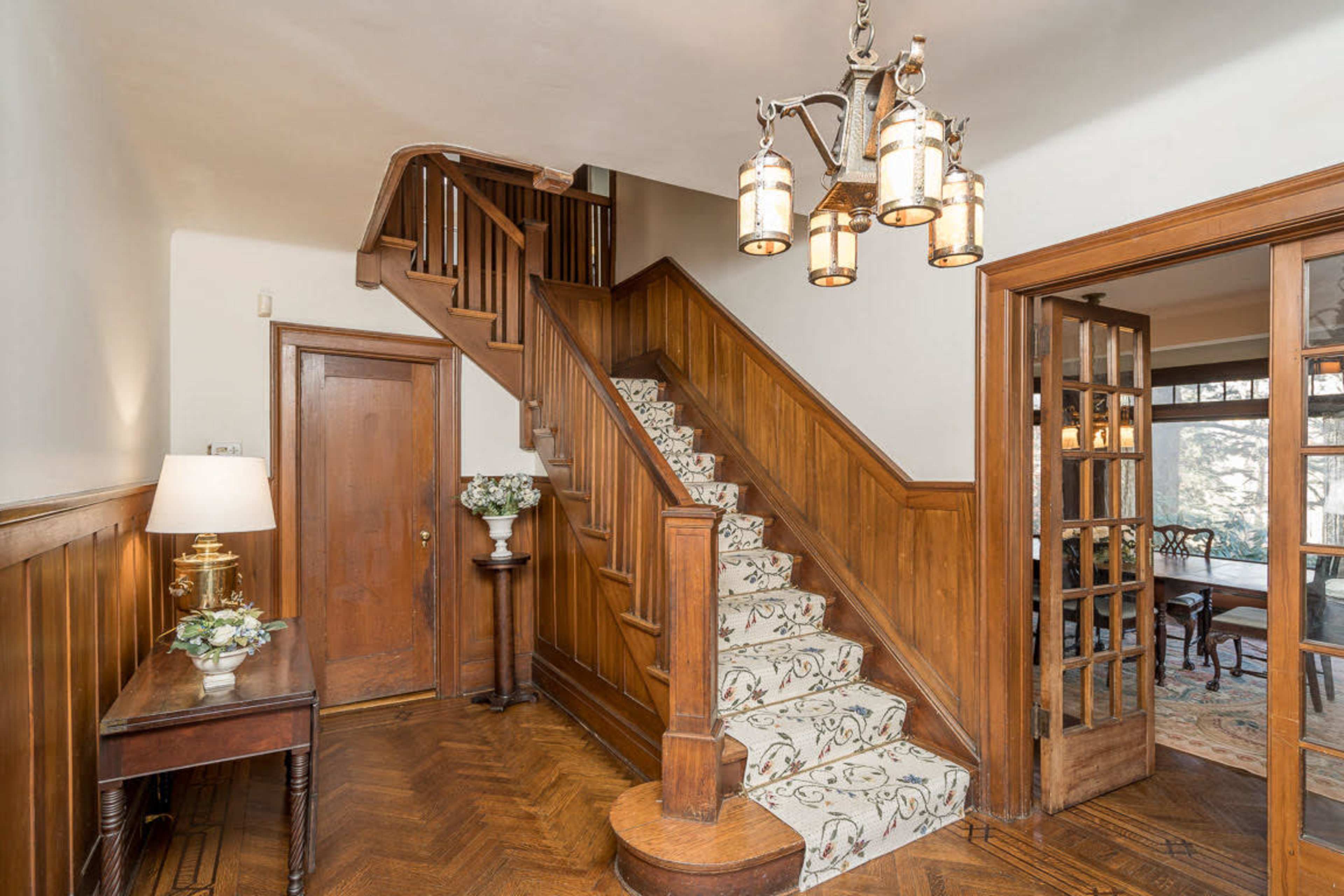 The image shows a wooden entryway with a staircase leading to an upper floor, featuring ornate light fixtures and decorative stairs.