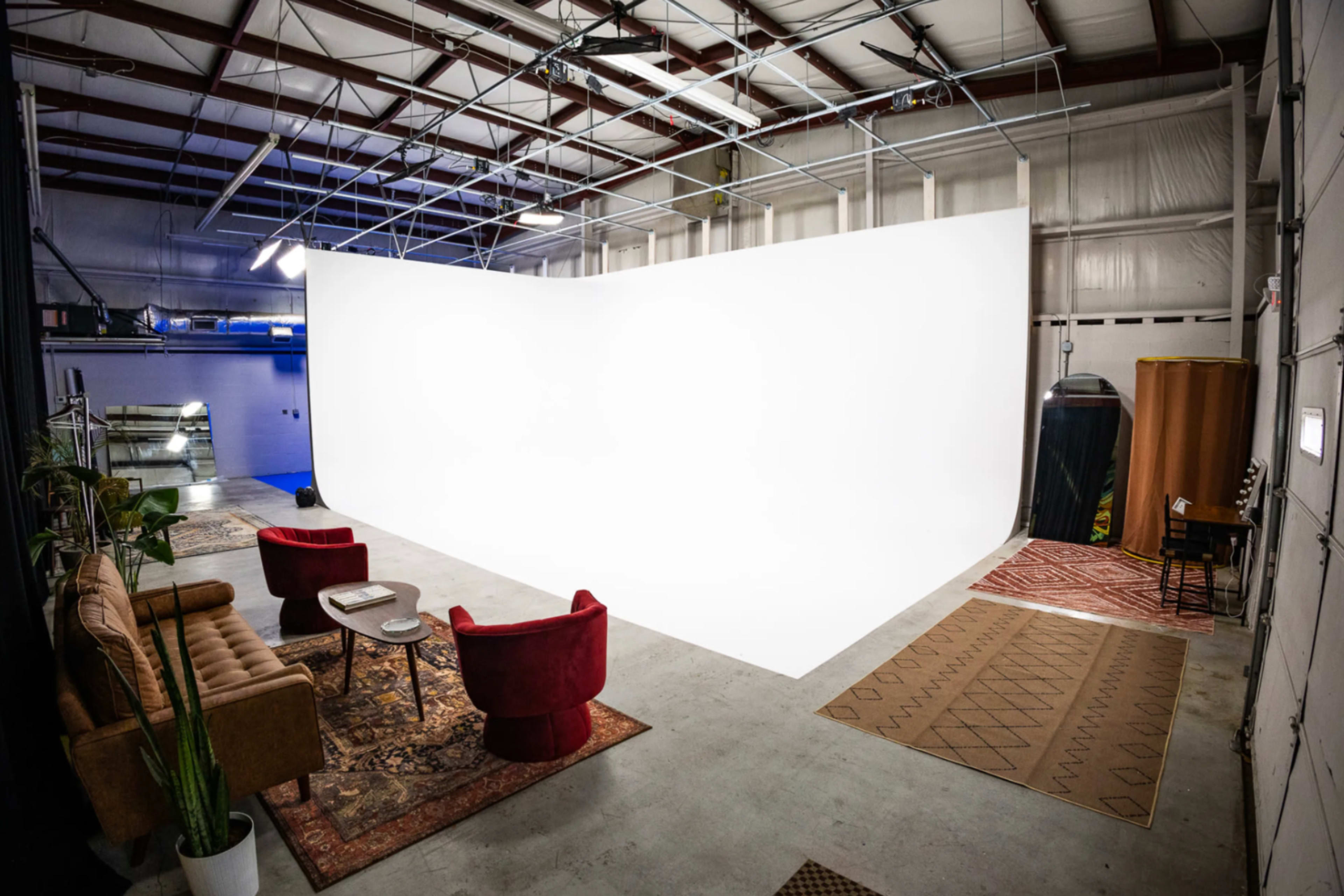 The image shows a spacious photography studio featuring a large white backdrop, a sofa, two red chairs, and decorative rugs.