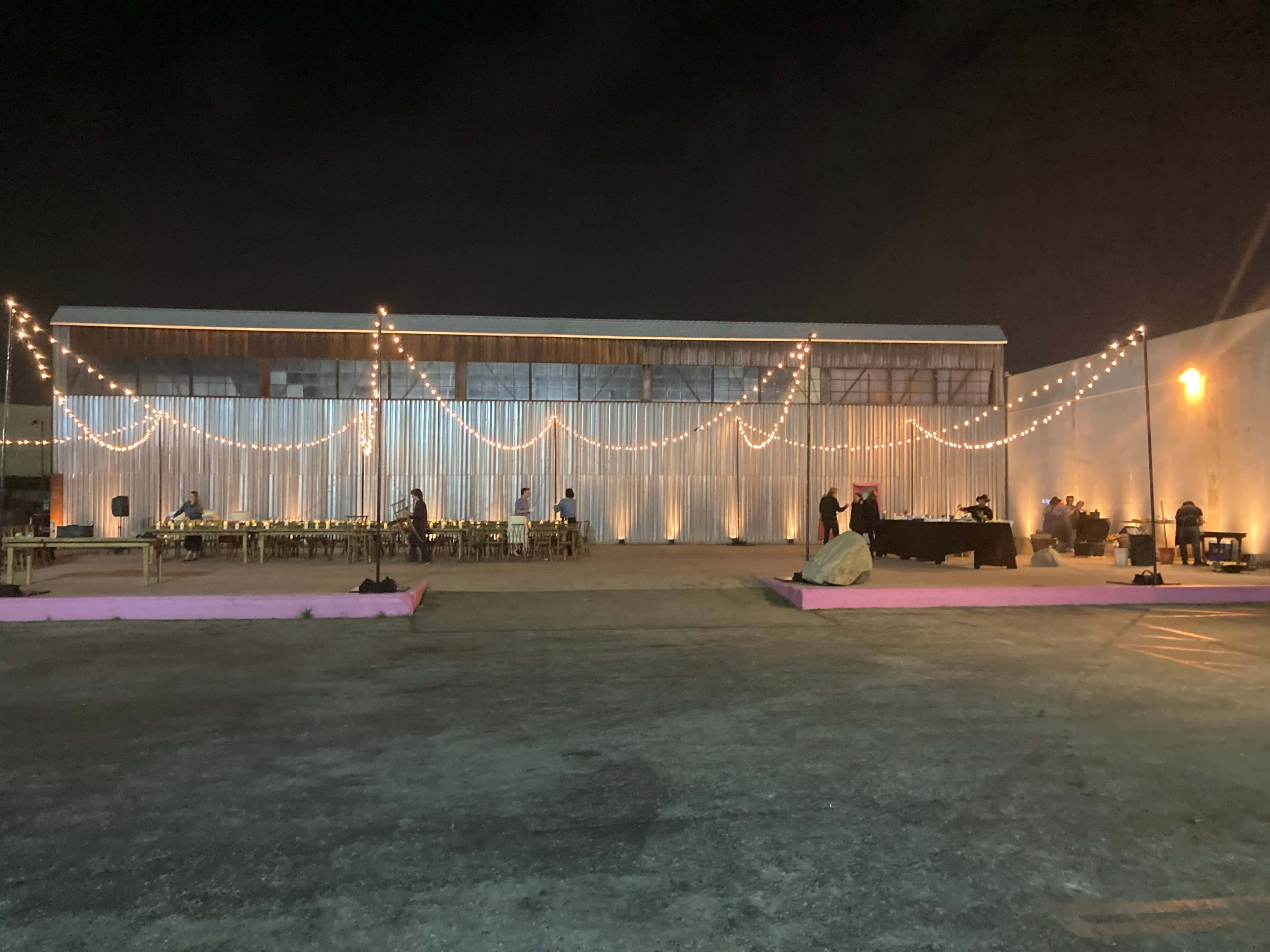 The image shows a large outdoor event space featuring string lights draped across a metal building, with tables set for a gathering and a bar area visible.