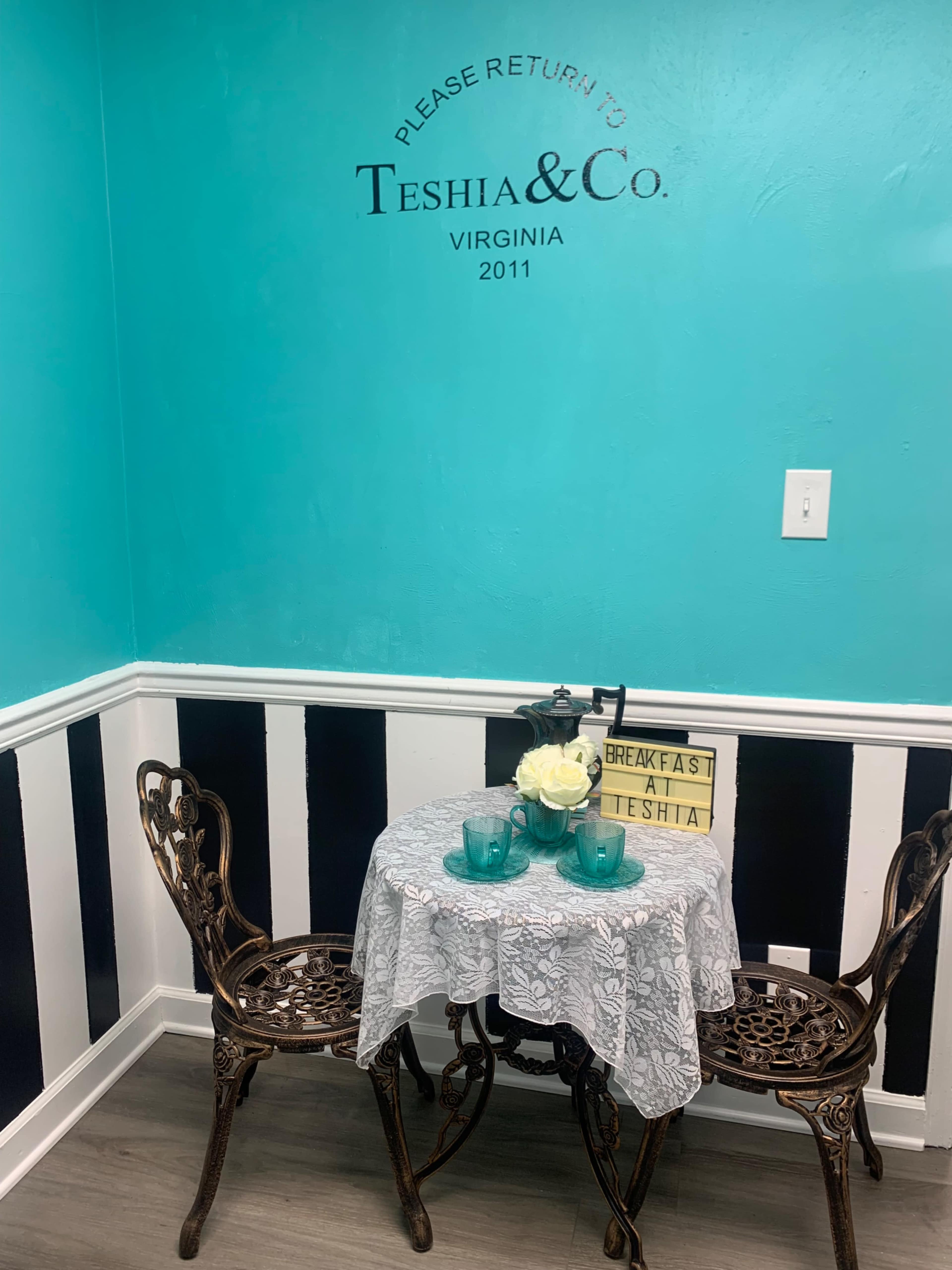 A small table set for two with a lace tablecloth, turquoise glasses, and a vase of white flowers is placed against a turquoise wall adorned with black and white striped accents.
