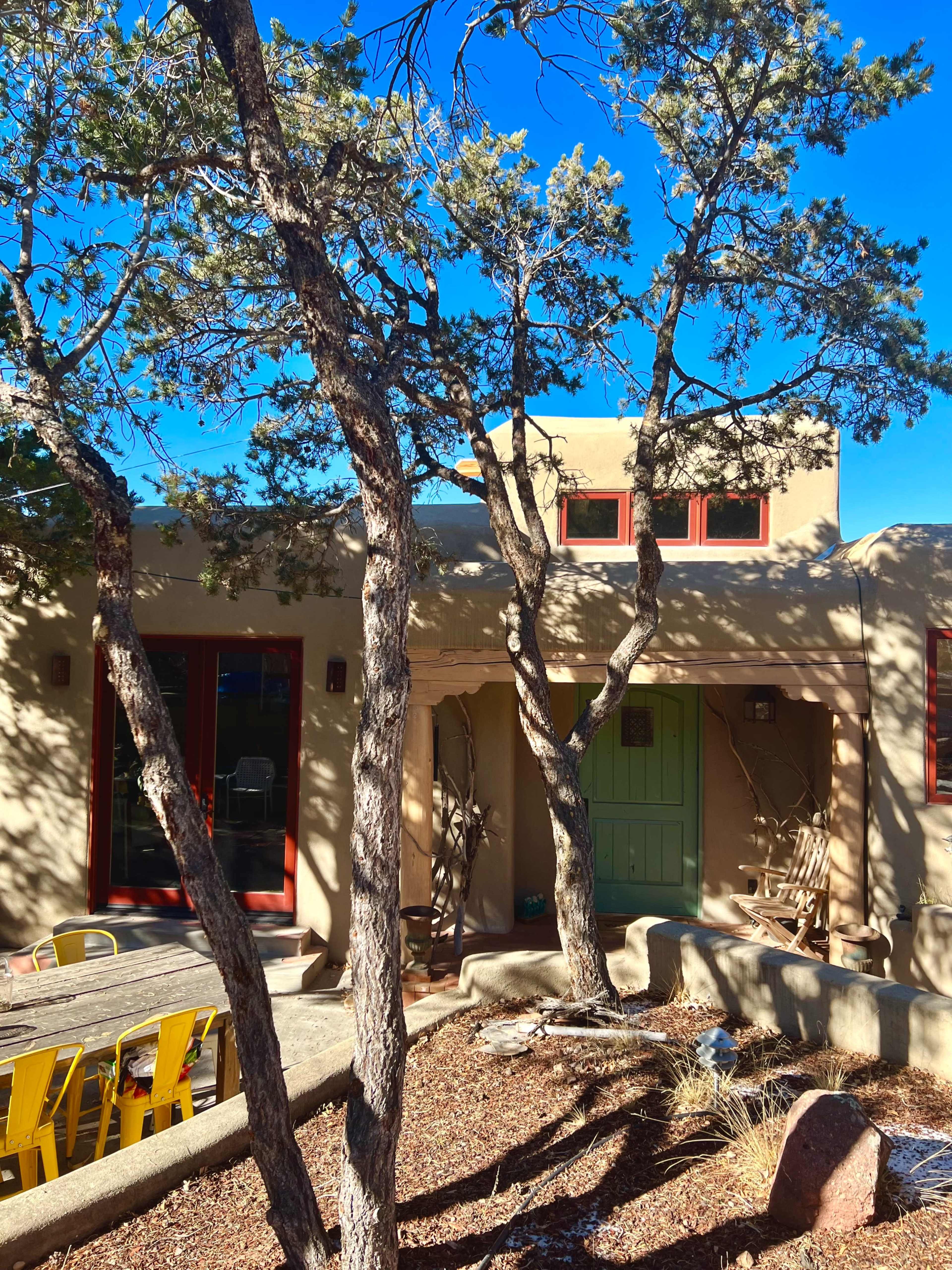 A southwestern-style adobe house with a green door and red window frames is surrounded by trees and a gravel landscape.