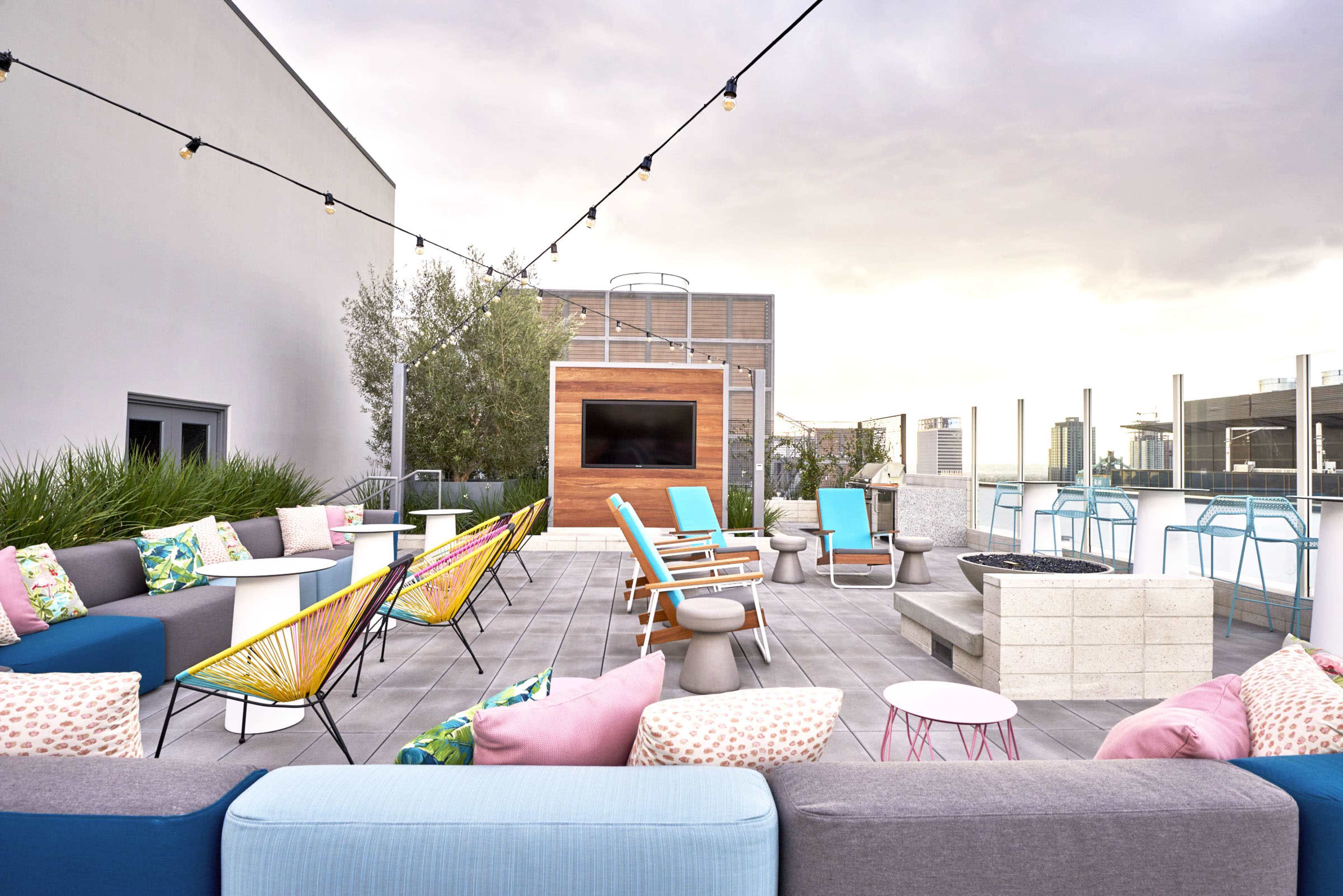 The image shows a modern rooftop lounge area with colorful seating, a TV, and a fire pit, surrounded by greenery and city views.