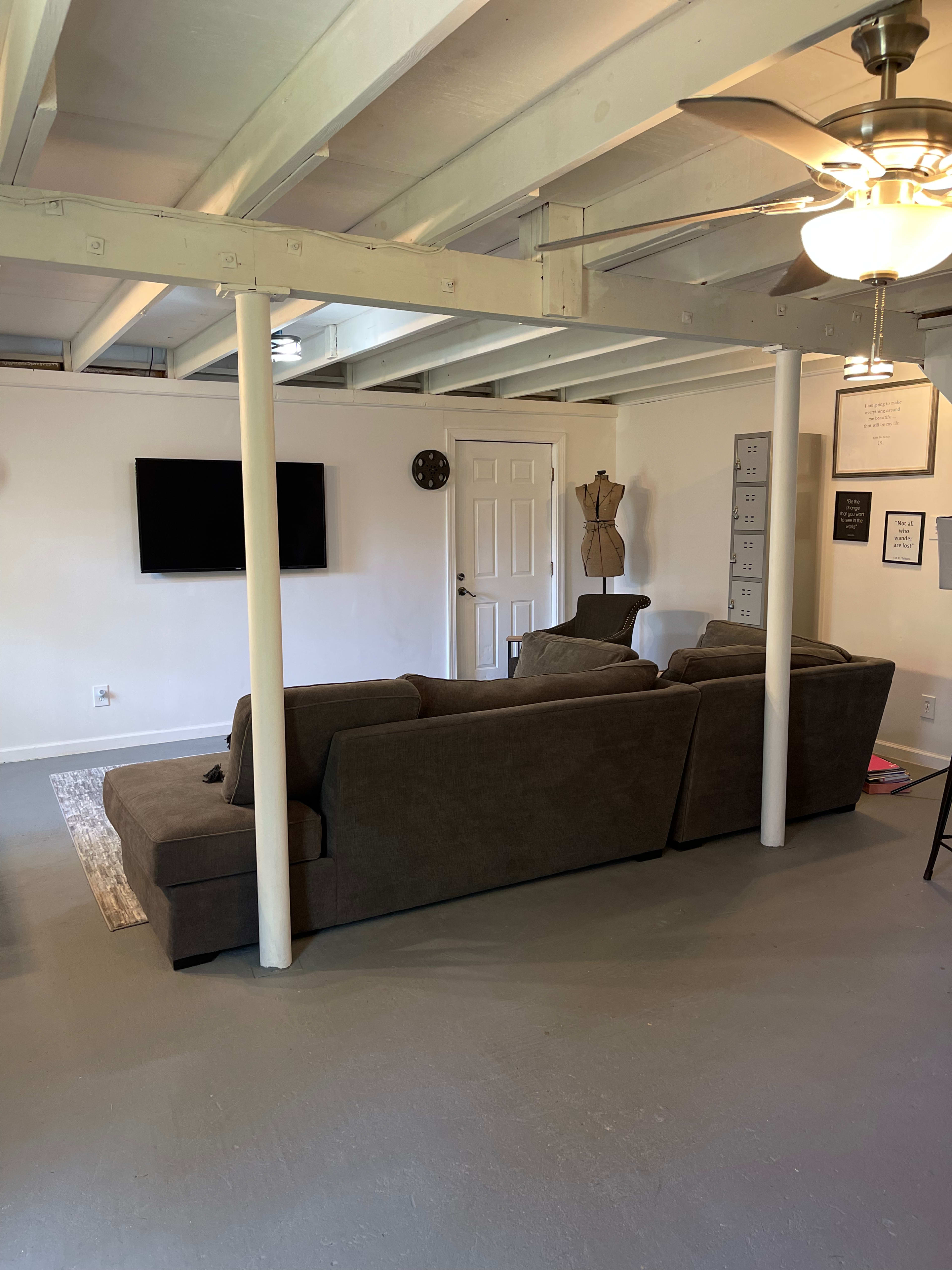 A living room features a sectional sofa, a wall-mounted TV, and a door leading outside, with white walls and exposed beams above.