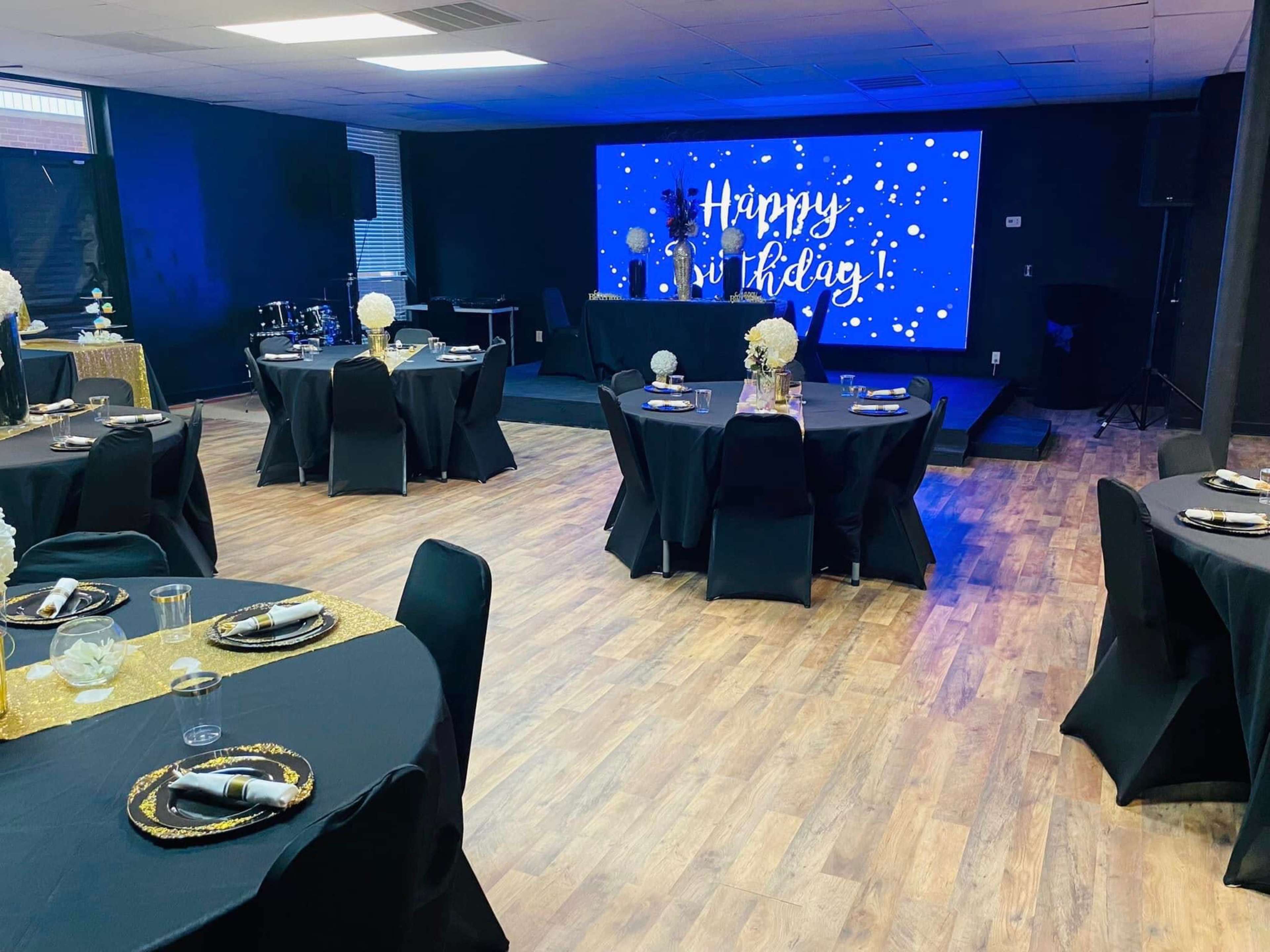 A decorated event space with black tables set for a celebration, a stage featuring a "Happy Birthday" display, and floral arrangements on the tables.